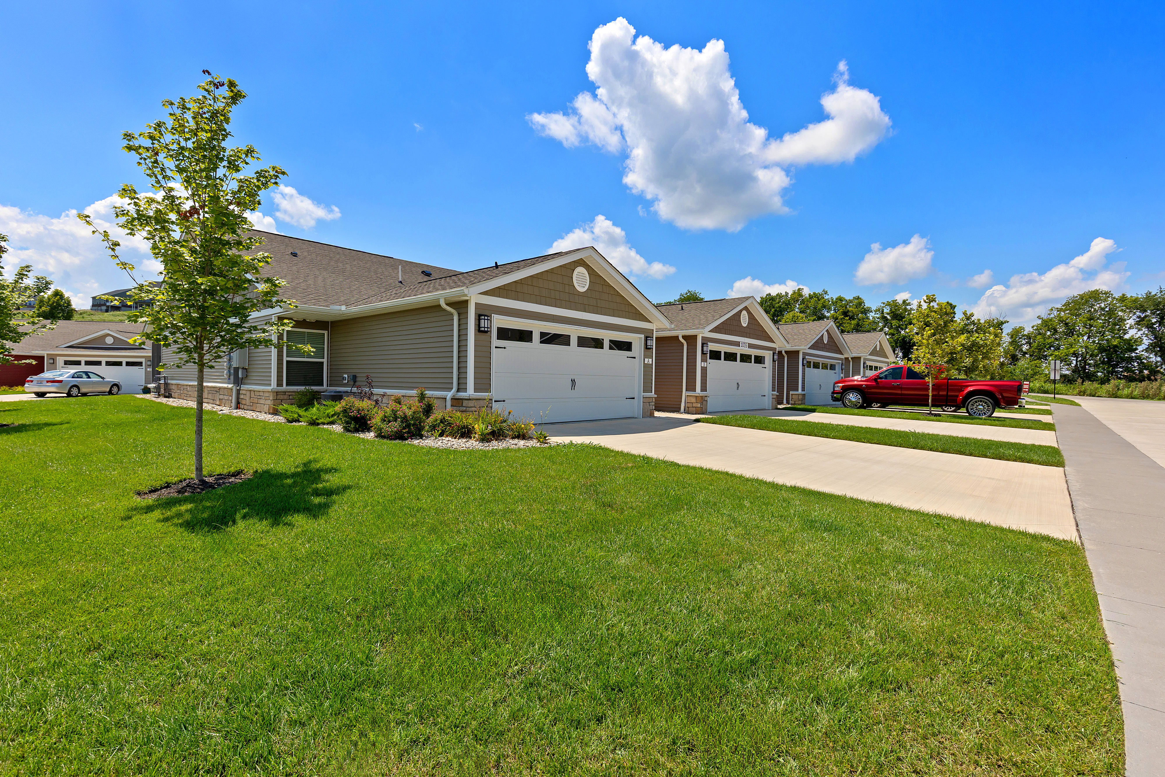 Row of single-story suburban houses with attached garages, driveways, green lawns, and a red pickup truck parked in front under a blue sky with scattered clouds.