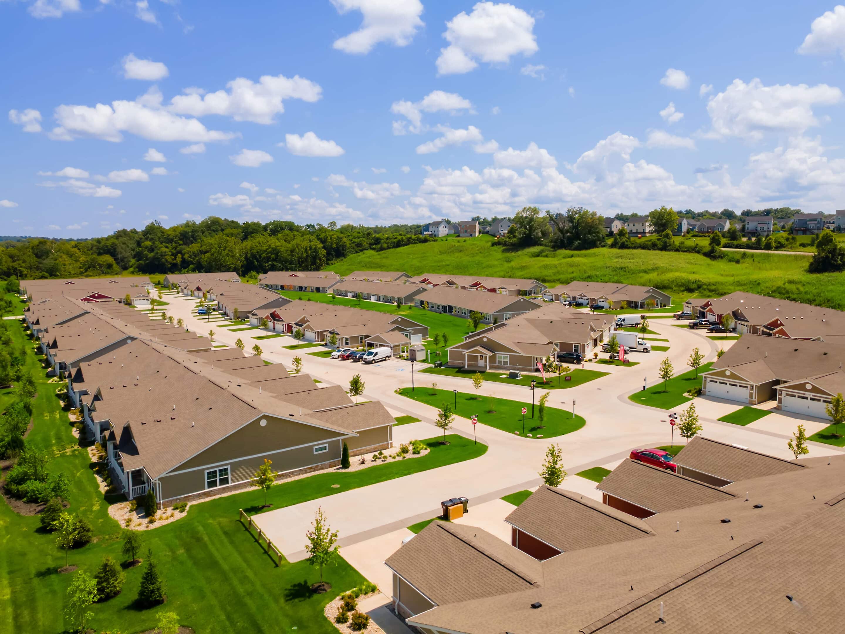 Aerial view of a suburban neighborhood with rows of single-story houses, green lawns, and curved streets under a blue sky with scattered clouds.