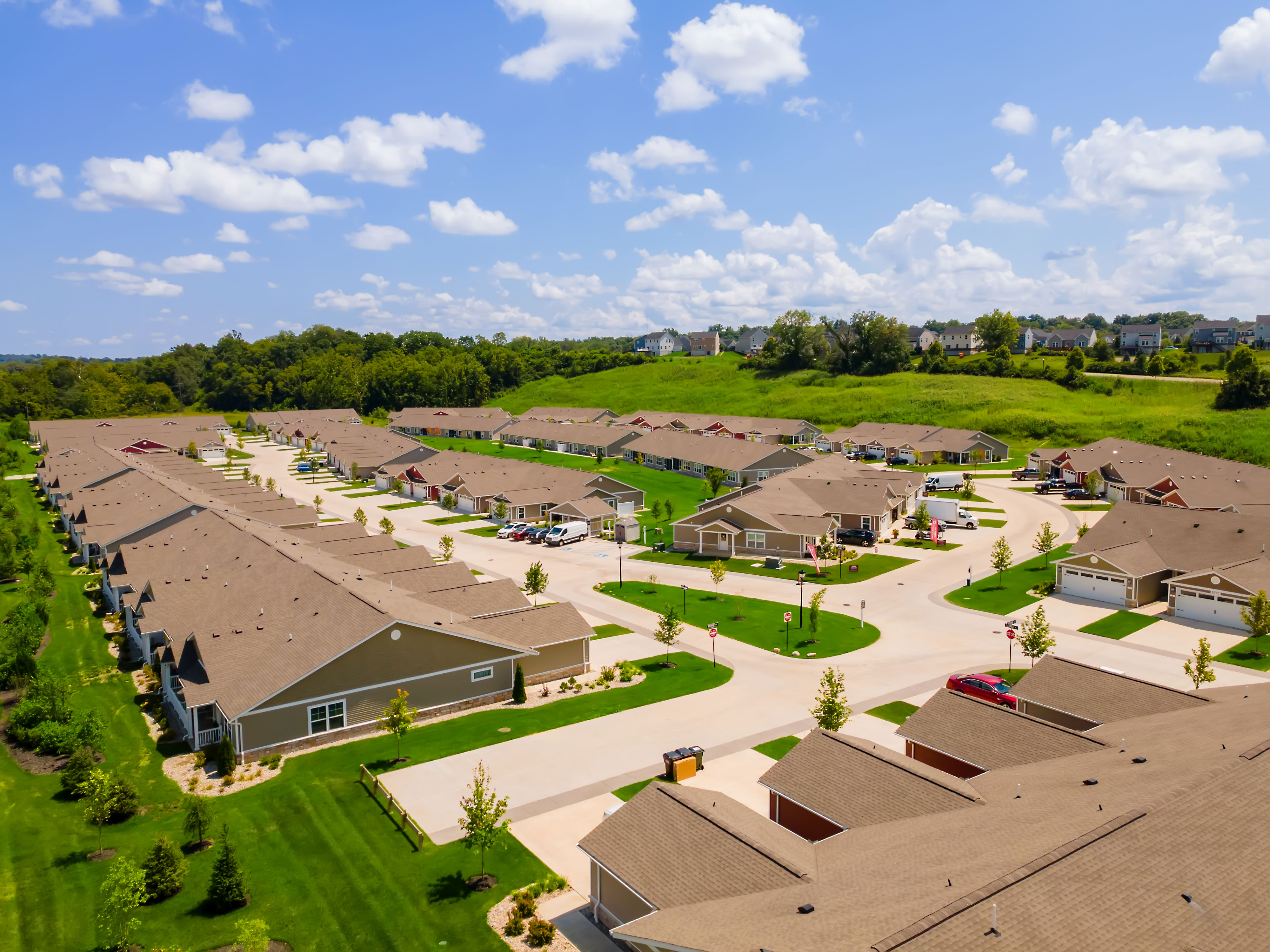 Aerial view of a suburban neighborhood with rows of single-story houses, green lawns, and curved streets under a blue sky with scattered clouds.