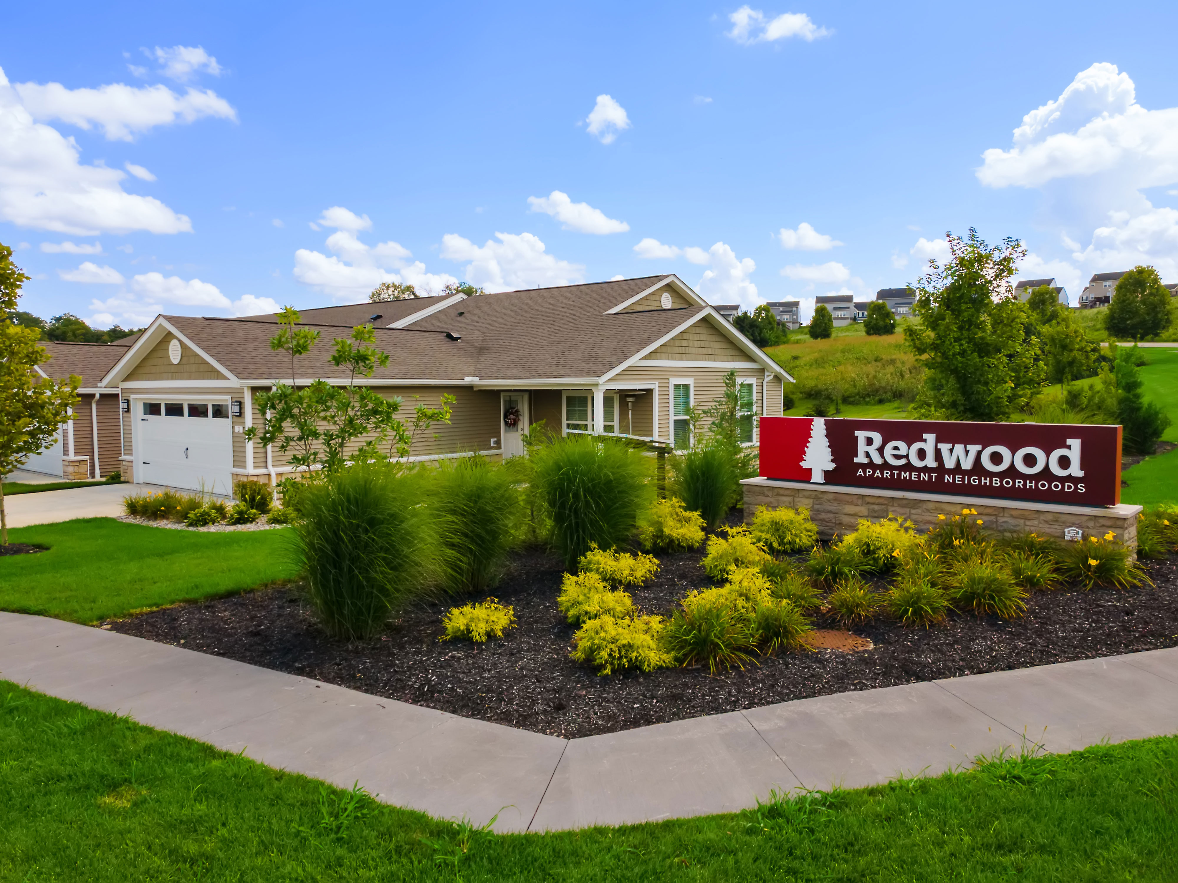 A single-story building with a garage sits behind a red sign reading "Redwood Apartment Neighborhoods," surrounded by landscaped greenery and a sidewalk under a blue sky.