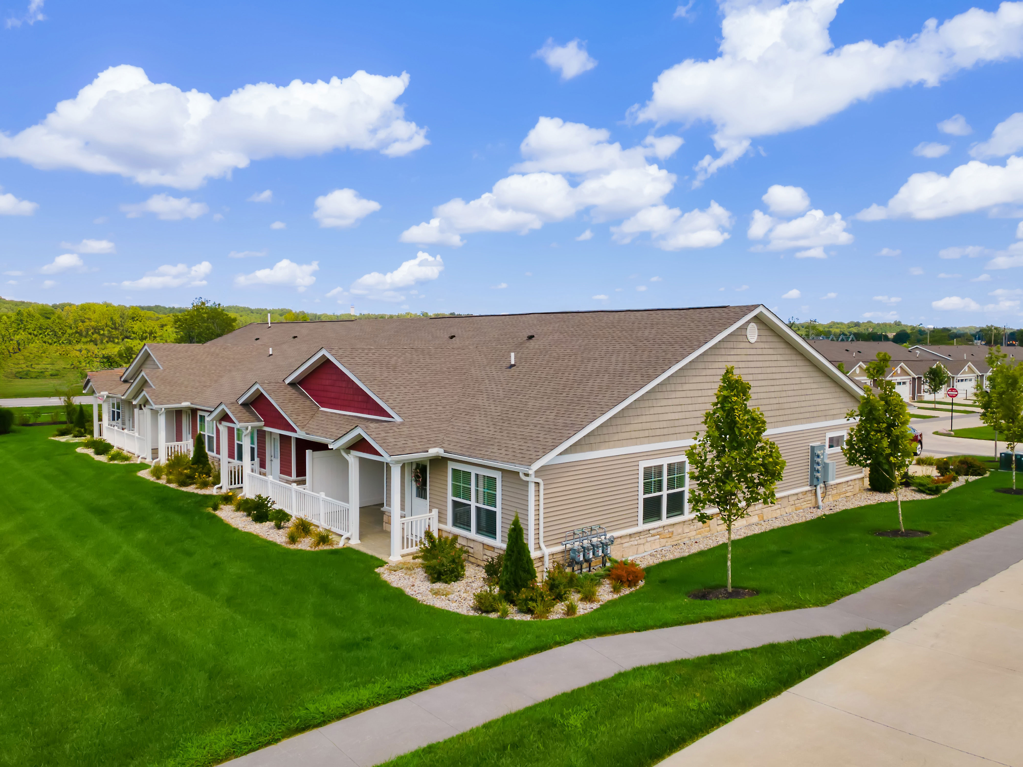 Single-story residential building with tan siding and red accents, surrounded by green lawns, young trees, a sidewalk, and a partly cloudy blue sky.