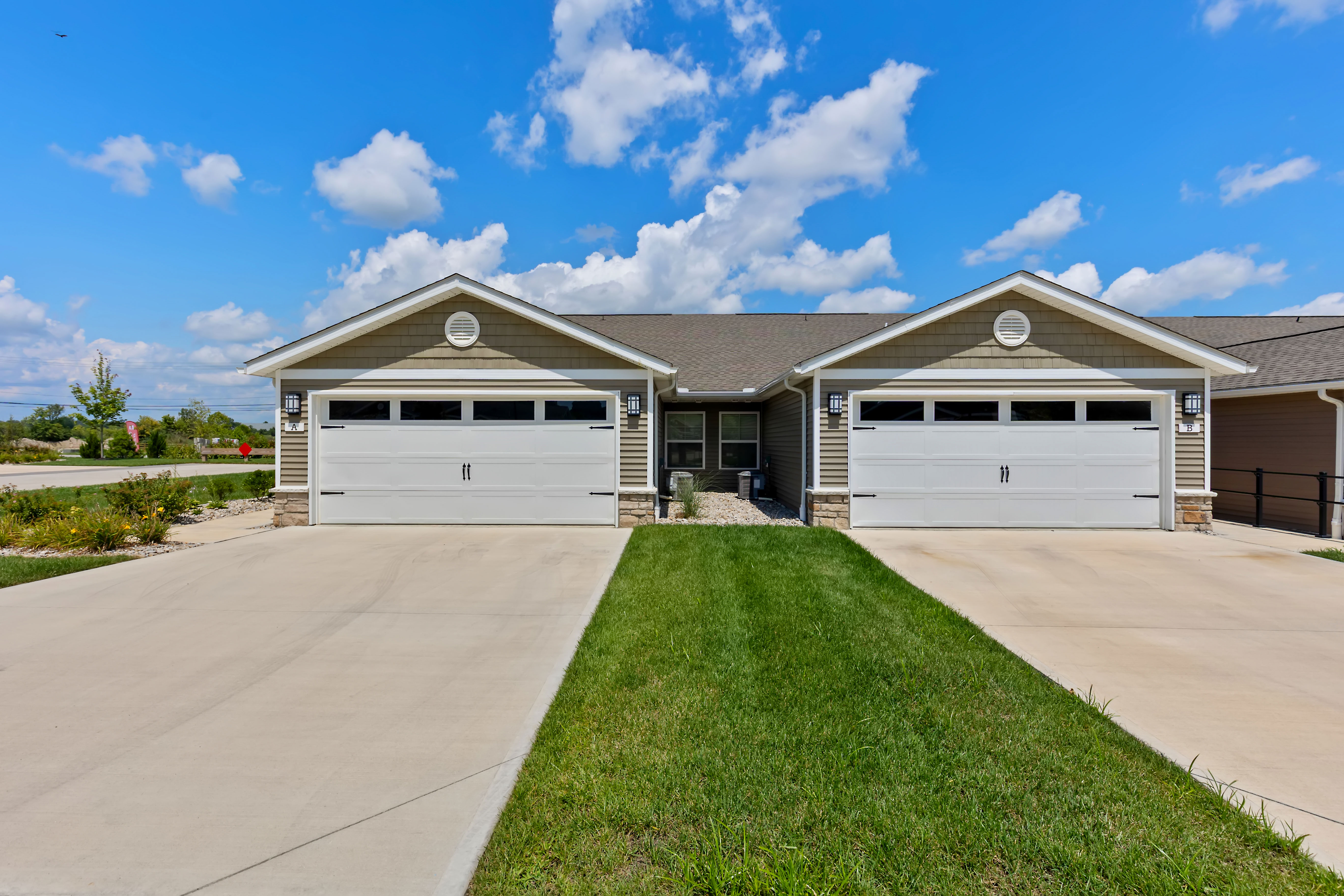 A duplex with two attached garages, beige siding, white garage doors, and a shared green lawn under a blue sky with scattered clouds.