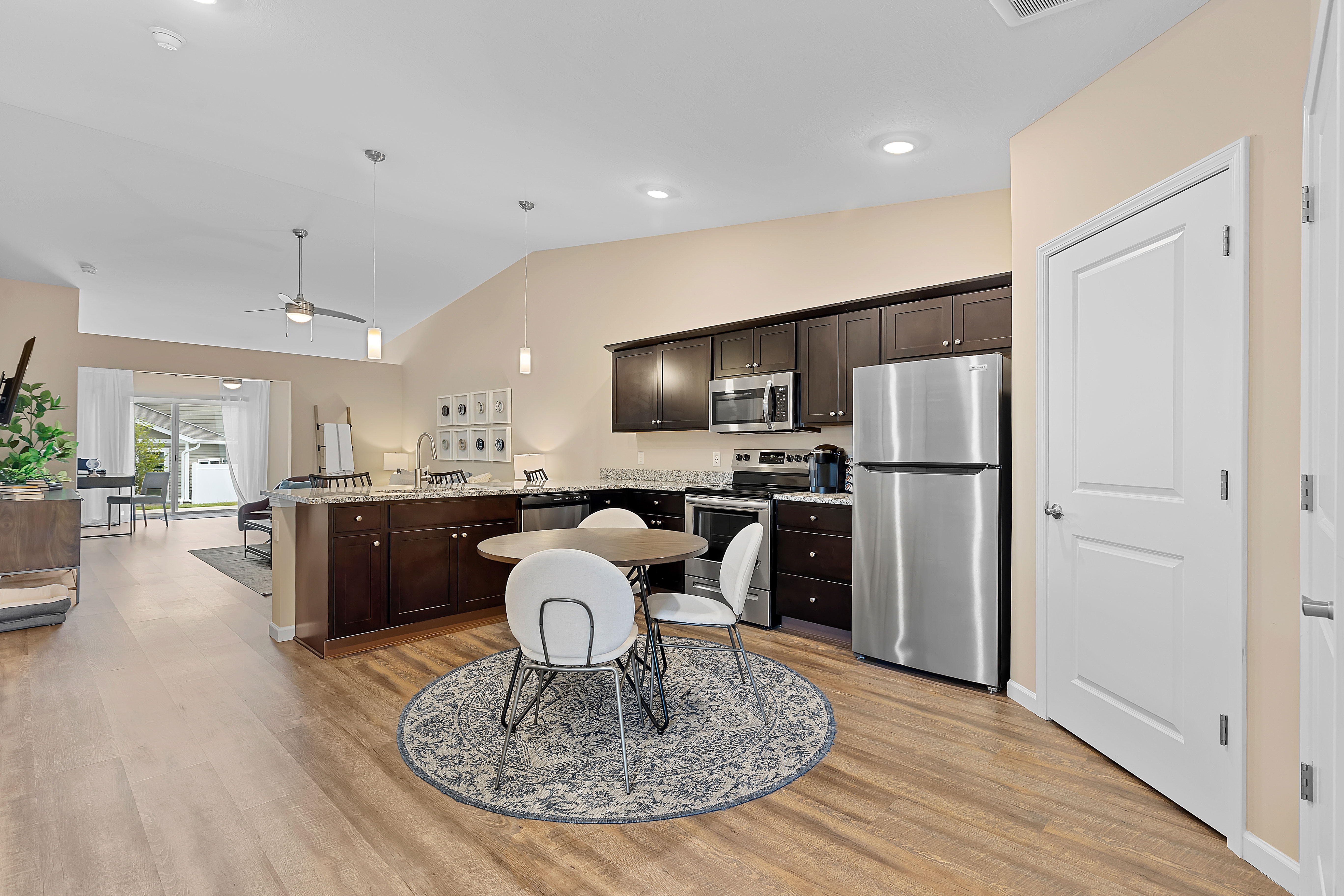 Modern kitchen with dark wood cabinets, stainless steel appliances, a round dining table with four chairs on a rug, and an open layout connecting to the living area.