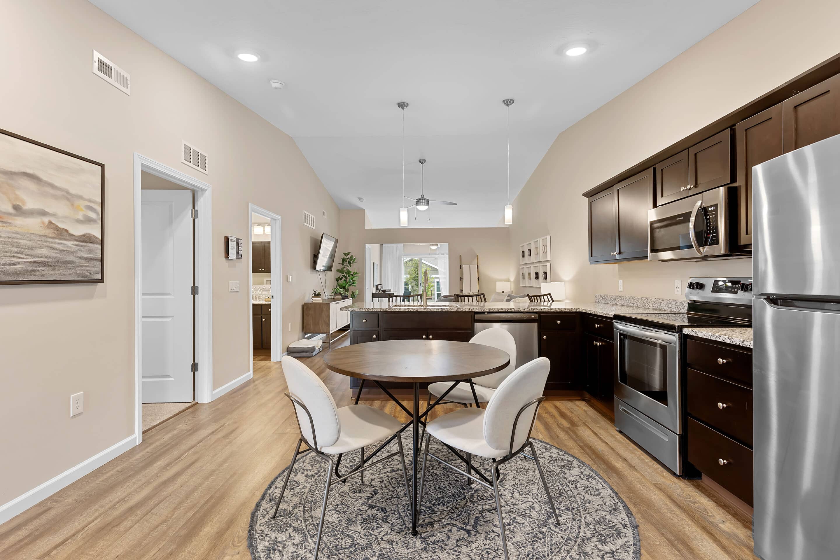 Modern kitchen and dining area with stainless steel appliances, dark cabinets, round table with four chairs on a rug, and wood flooring under bright ceiling lights.