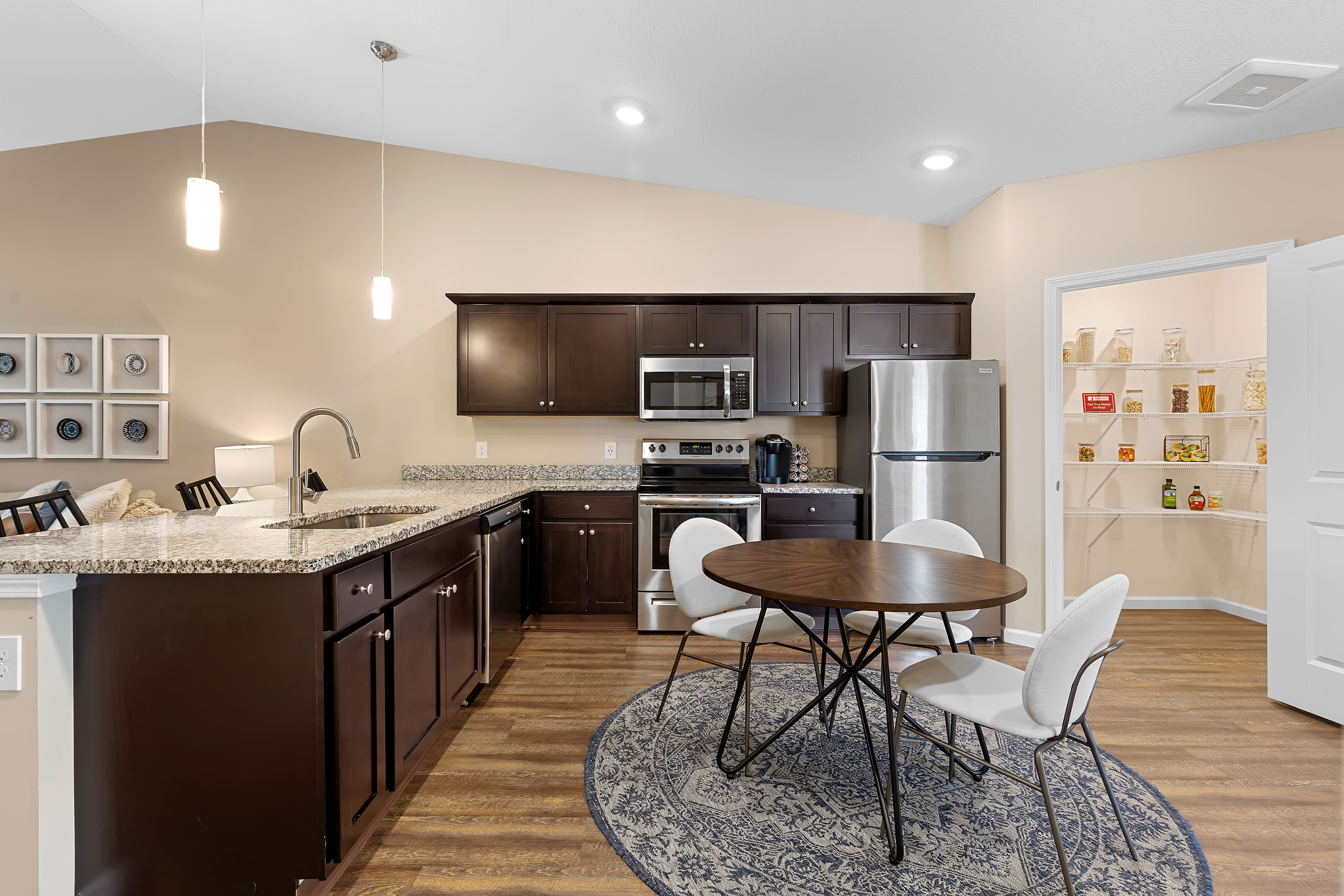 Modern kitchen with dark wood cabinets, stainless steel appliances, granite countertops, round dining table with four chairs, and an open pantry stocked with food items.