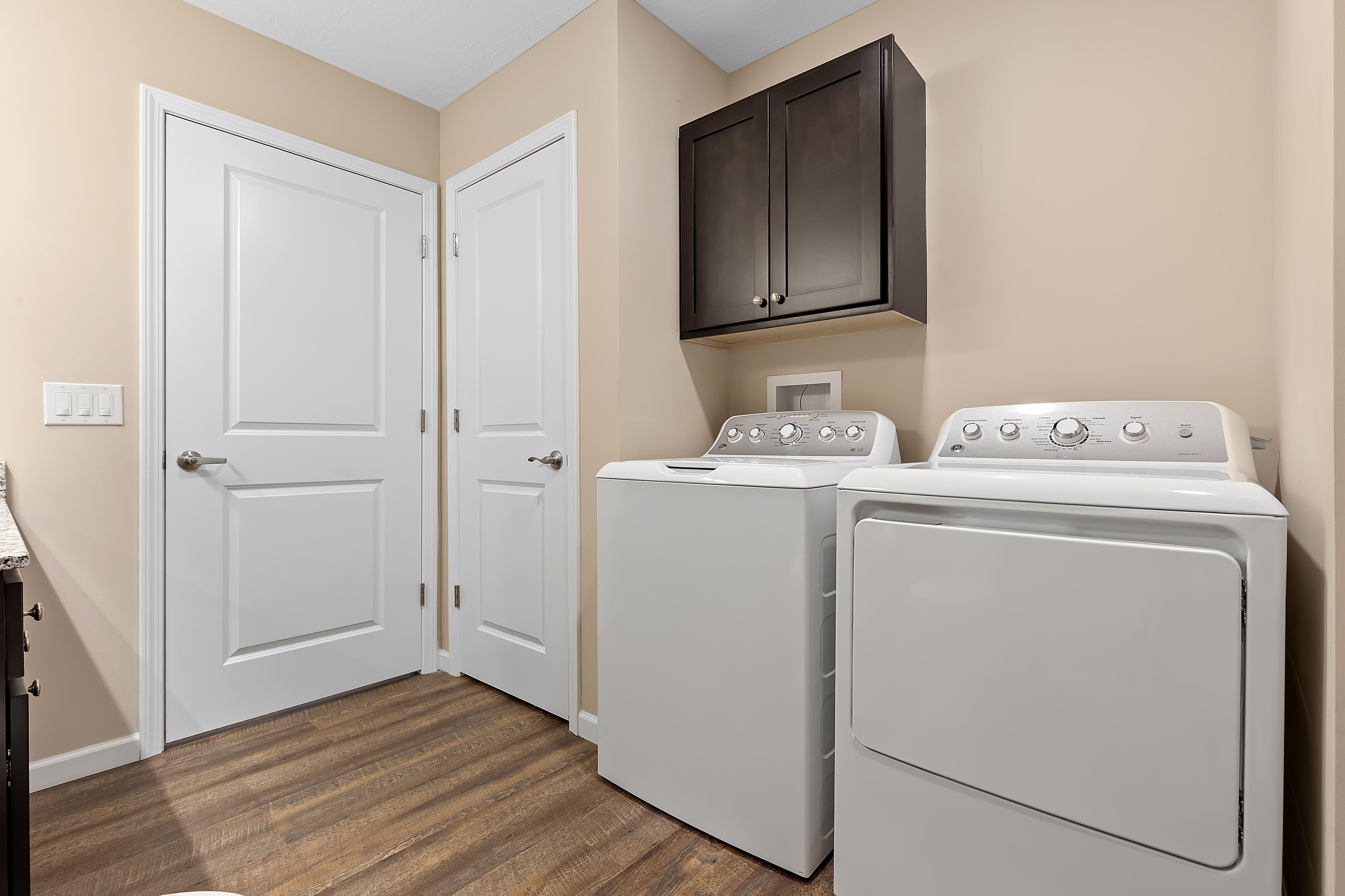 Laundry room with a washing machine and dryer side by side, dark wood cabinet above, beige walls, wood-look flooring, and two closed white doors.