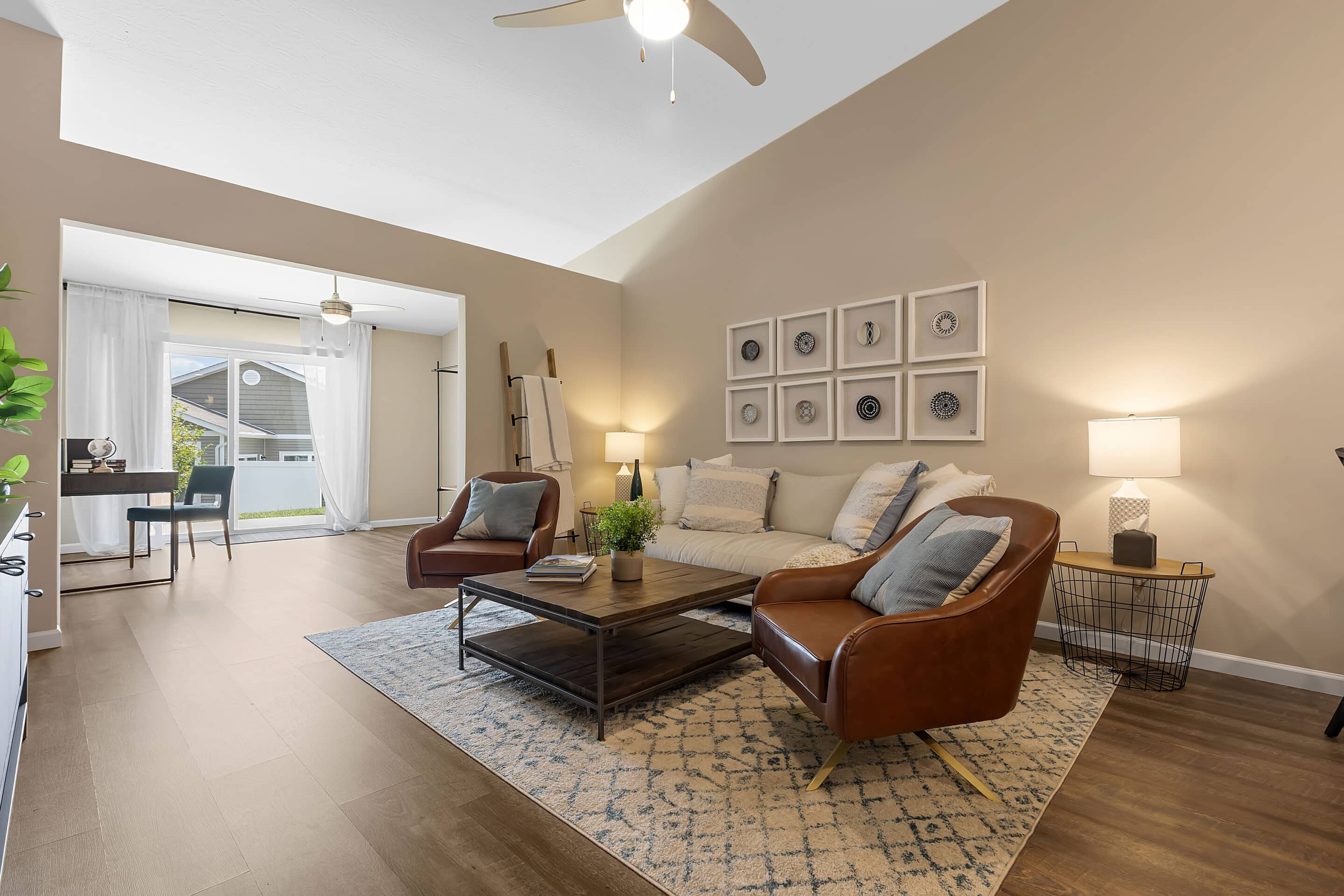 A modern living room with a beige sofa, two brown leather chairs, a wooden coffee table, wall art, and a view into a bright home office with large window and desk.