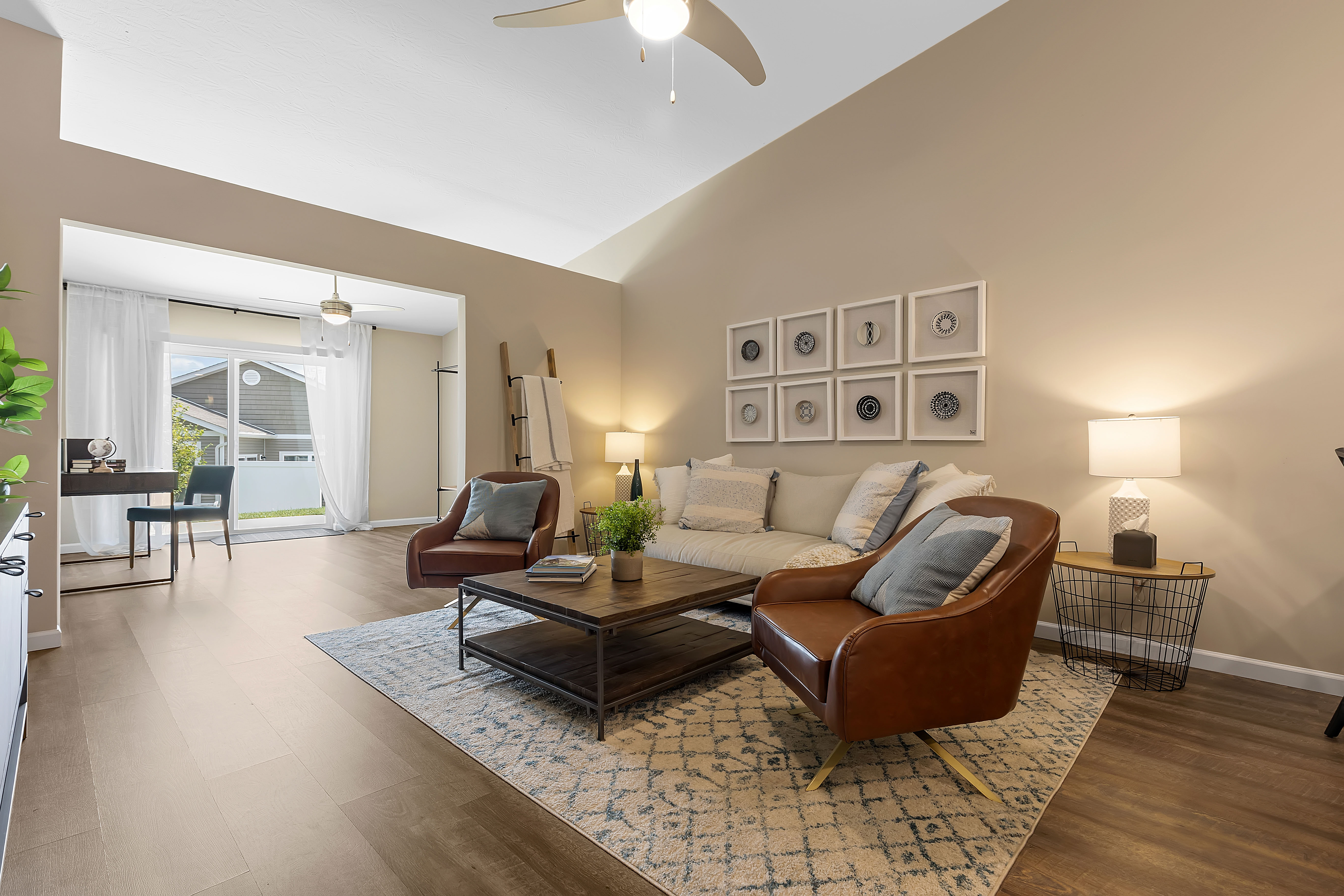 A modern living room with a beige sofa, two brown leather chairs, a wooden coffee table, wall art, and a view into a bright home office with large window and desk.