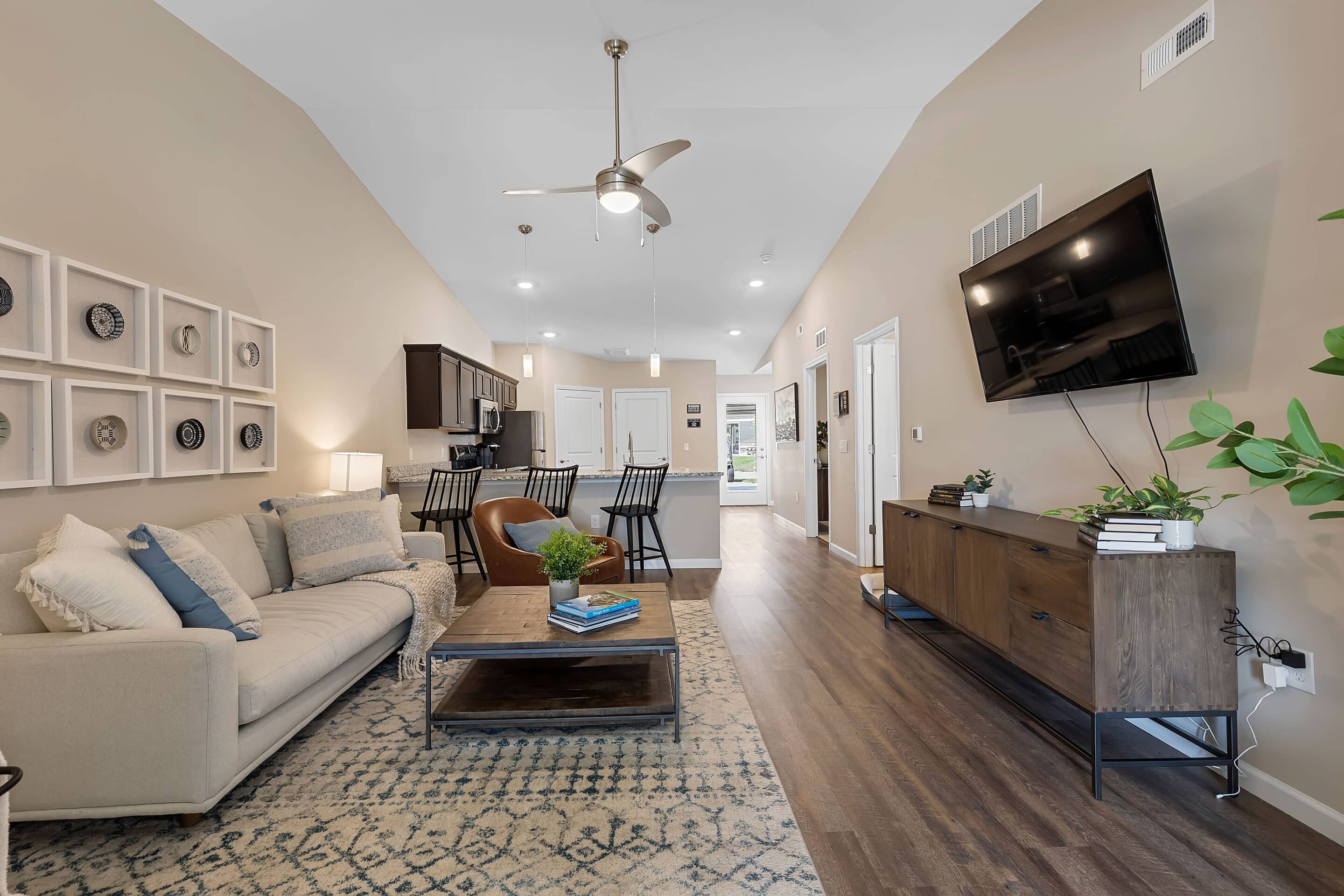 Modern living room with beige sofa, patterned rug, coffee table, wall-mounted TV, and open kitchen with bar stools in the background. Decor includes plants and framed artwork.