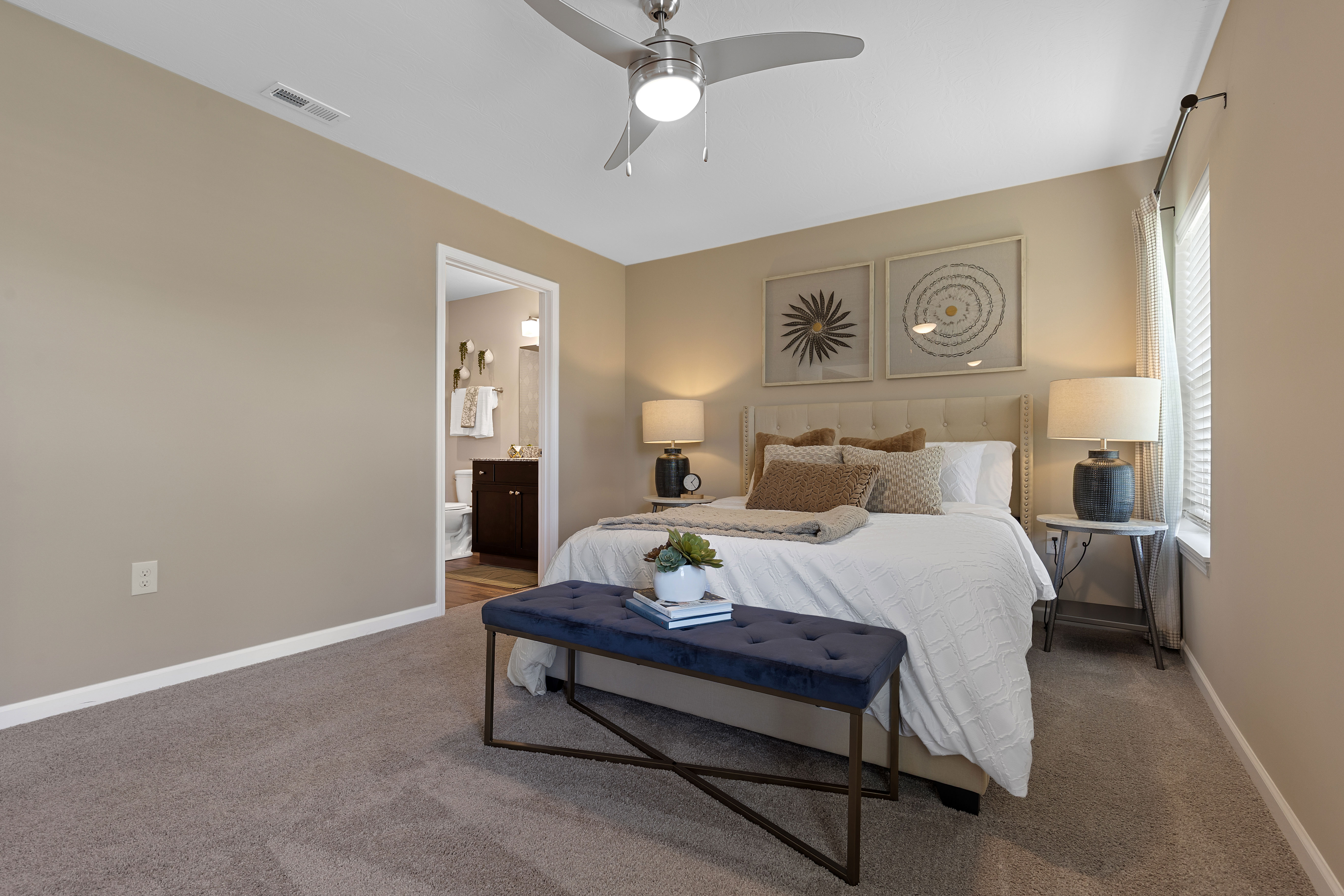 A modern bedroom with beige walls, a bed with white bedding, two side tables with lamps, a blue bench at the foot of the bed, and wall art above the headboard.