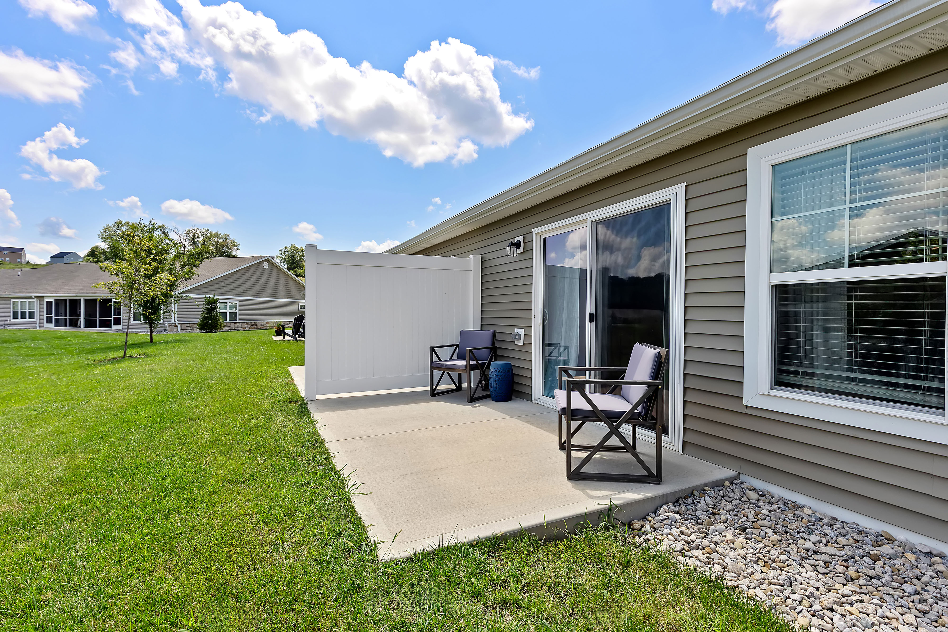 A small concrete patio with two chairs and a privacy screen outside a single-story house with beige siding and sliding glass doors, surrounded by grass and neighboring homes.