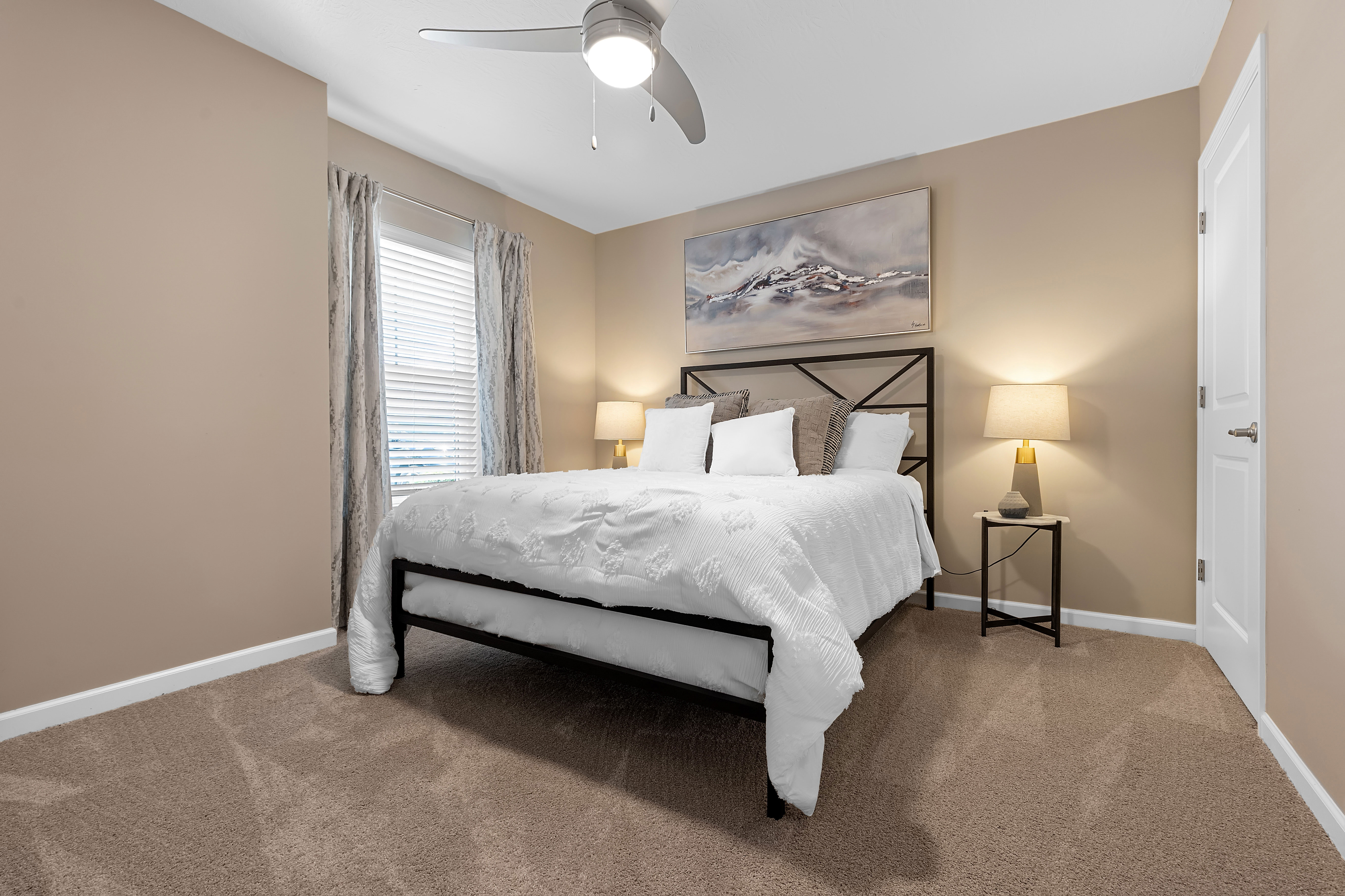 A neatly made bed with white bedding is centered in a beige bedroom, flanked by two nightstands with lamps, and a landscape painting hangs above the headboard.