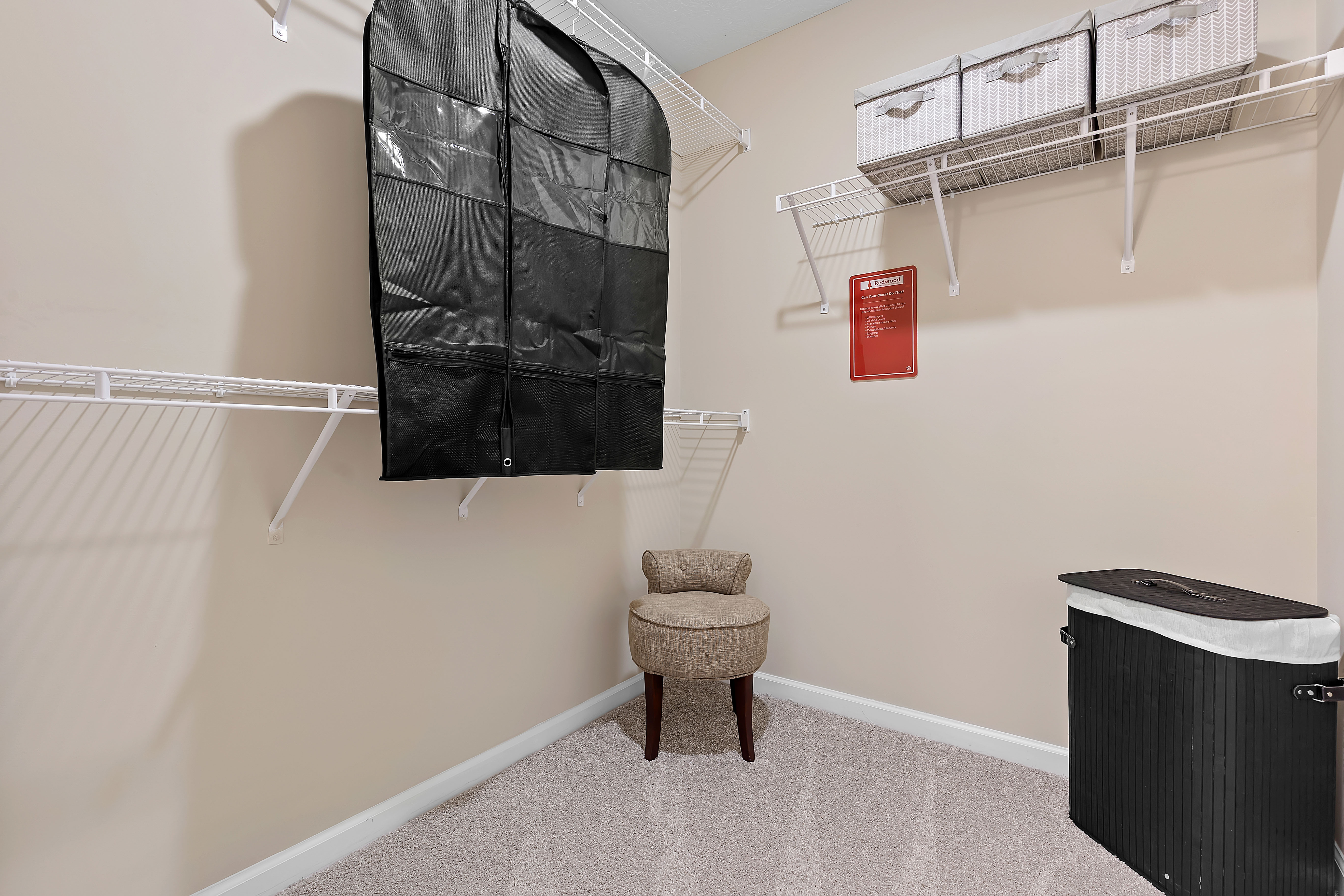 Walk-in closet with wire shelves, storage bins, a black garment bag, a beige upholstered chair, and a black laundry hamper on carpeted flooring.