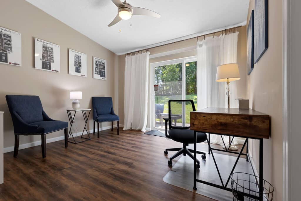 Home office with wood desk, black chair, two blue armchairs, side tables with lamps, wall art, large window with sheer curtains, and view of green backyard.