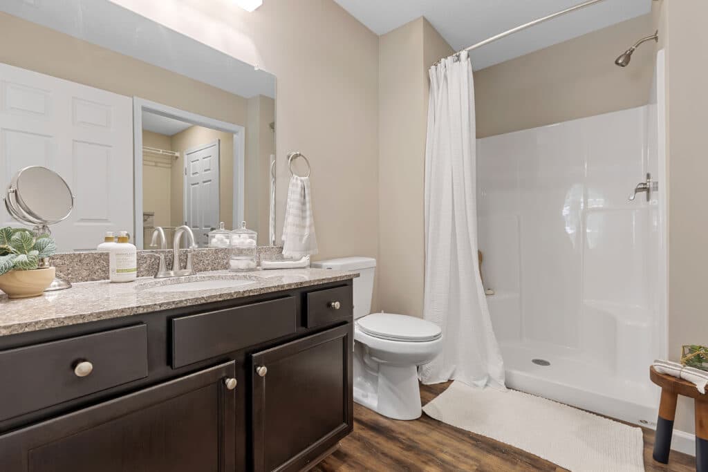 Modern bathroom with dark wood vanity, granite countertop, white sink, toilet, glass soap dispenser, towel, and a walk-in shower with a white curtain on a curved rod.
