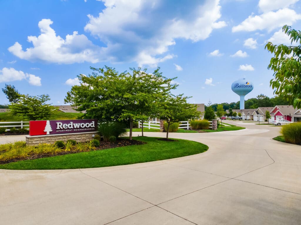 Entrance to Redwood Apartment Neighborhood with a curved driveway, landscaped greenery, and a Bellbrook water tower visible in the background under a partly cloudy sky.