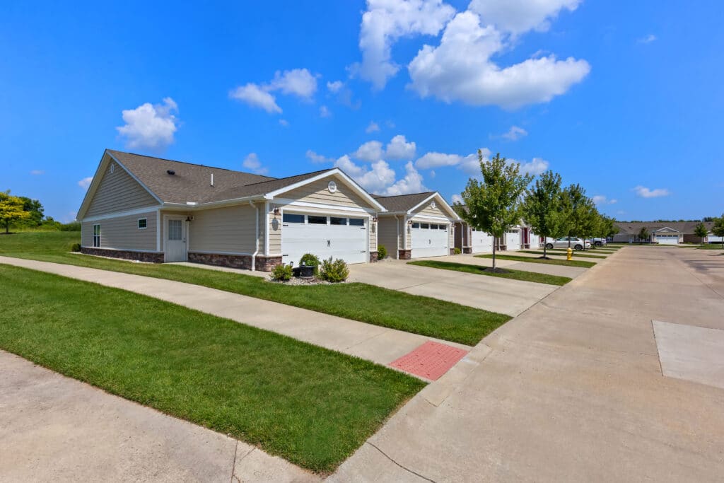 Row of modern single-story houses with attached garages, well-kept lawns, and a clear blue sky overhead. Concrete driveways and sidewalks run alongside the homes.