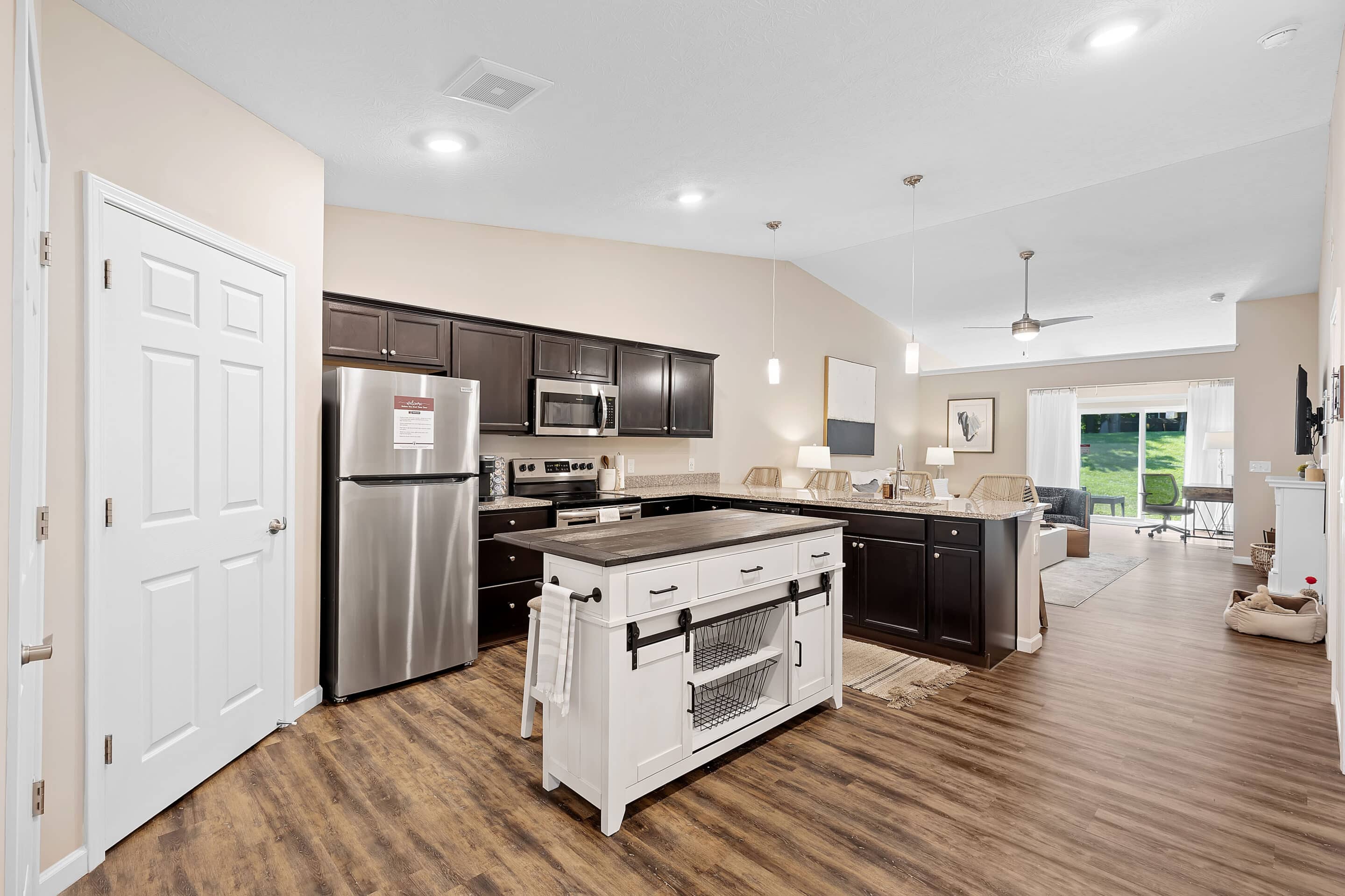 Modern open-concept kitchen and living room with stainless steel appliances, dark cabinets, island, and wood flooring, leading to a bright dining area and patio doors.