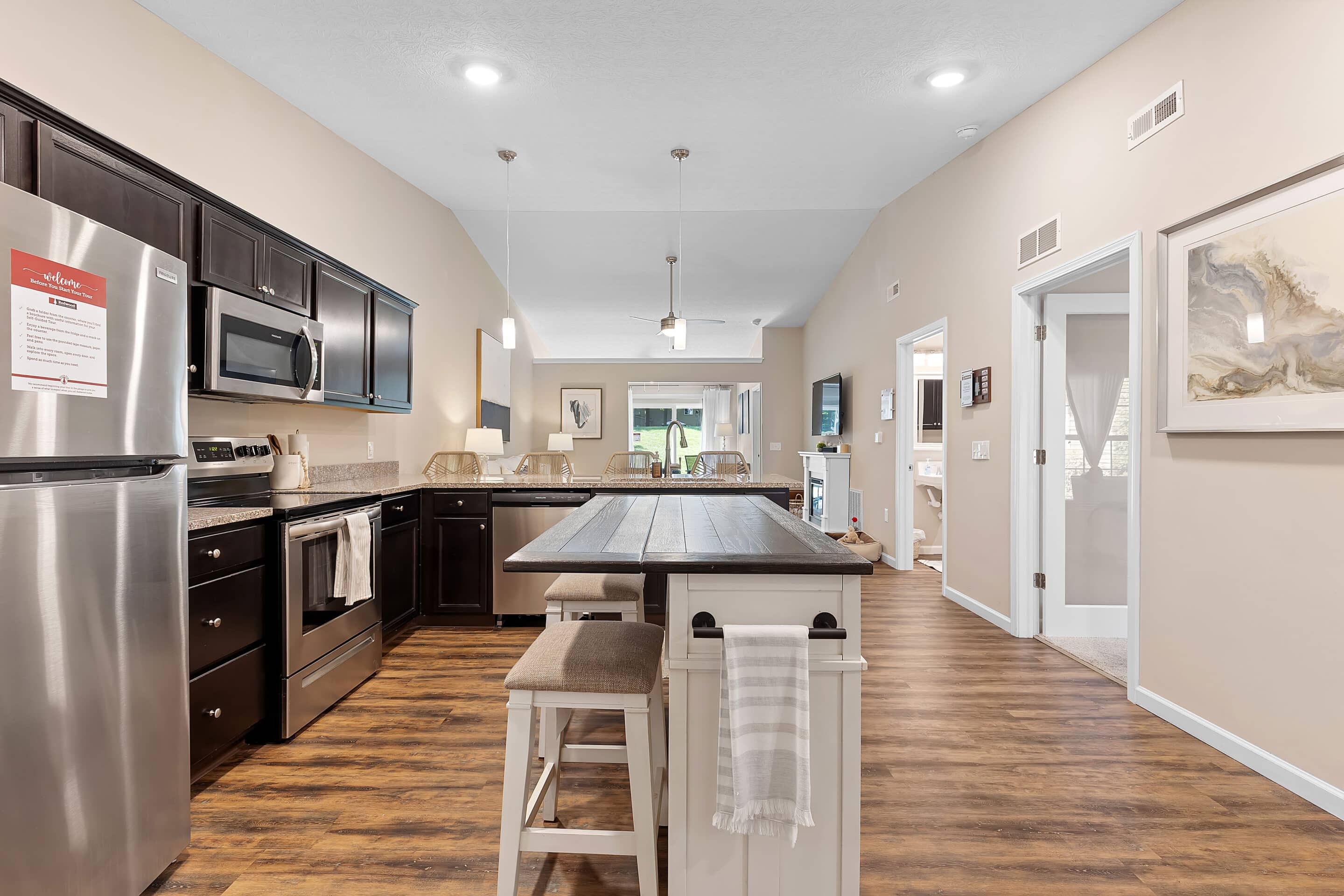 Modern kitchen with dark cabinets, stainless steel appliances, a center island with seating, and view into a living area; wood-style flooring throughout.