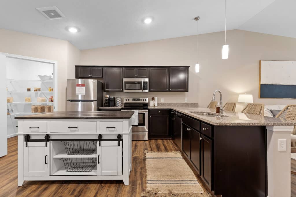 Modern kitchen with dark cabinets, stainless steel appliances, a white island with storage baskets, wood flooring, and pendant lights. A pantry is visible through an open door.