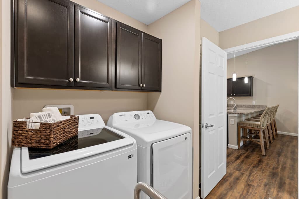 Laundry room with a washing machine and dryer beneath dark cabinets, a basket with towels on top, and an open door leading to a dining area with a table and chairs.