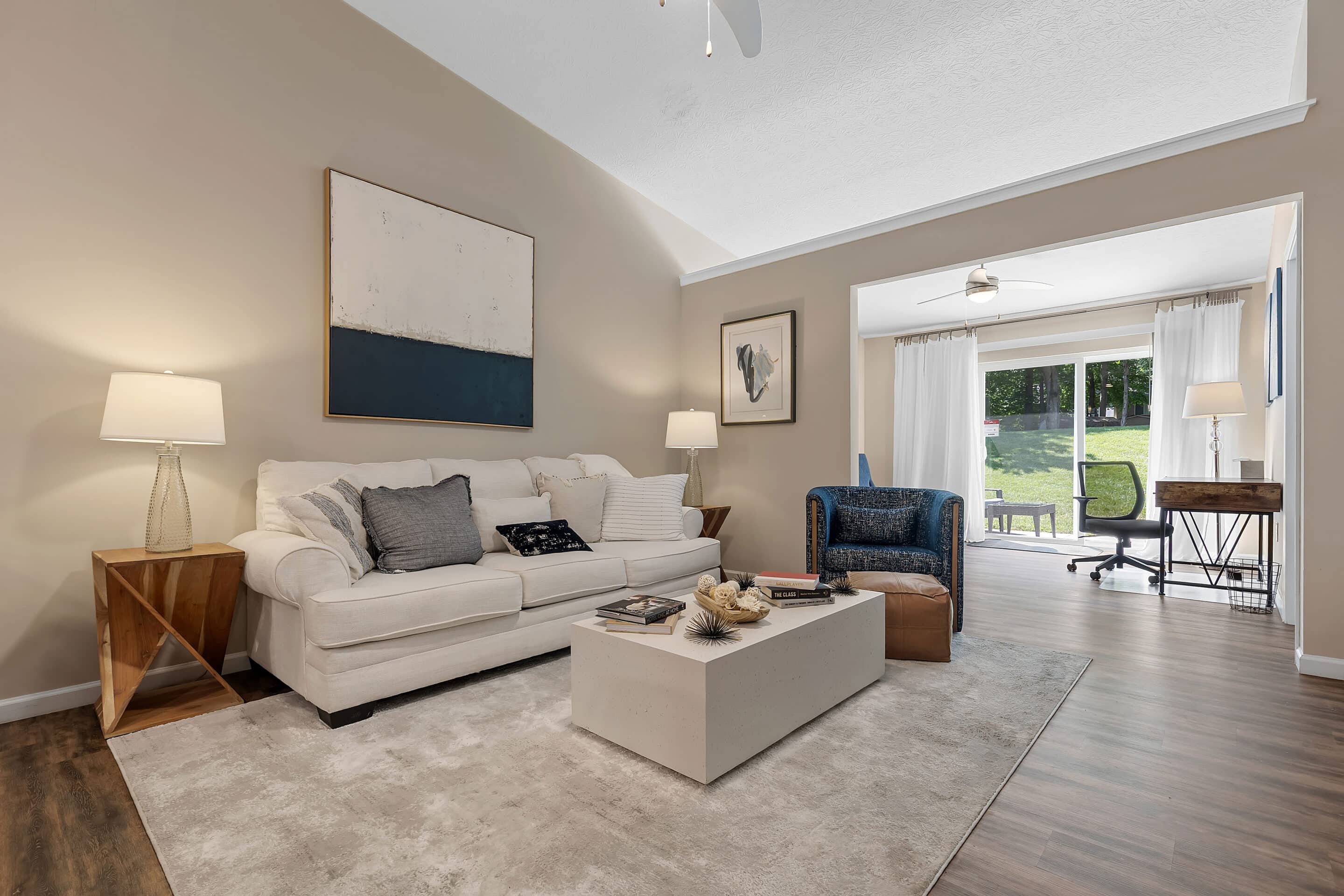 Modern living room with a white sofa, blue armchair, abstract art, coffee table, and a view into a sunlit office area with desk and sliding glass doors to the backyard.