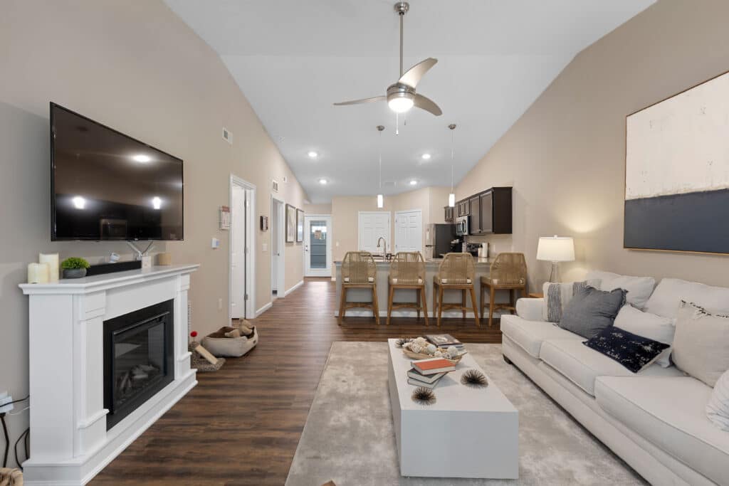 Modern open-concept living room and kitchen with a beige sofa, wall-mounted TV above a fireplace, island seating, hardwood floors, and neutral decor.