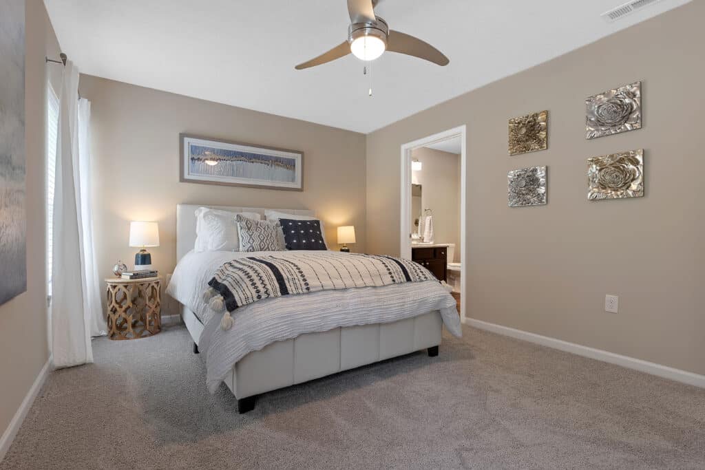 A bedroom with a neatly made bed, two bedside tables with lamps, a ceiling fan, wall art, and an open door leading to a bathroom.