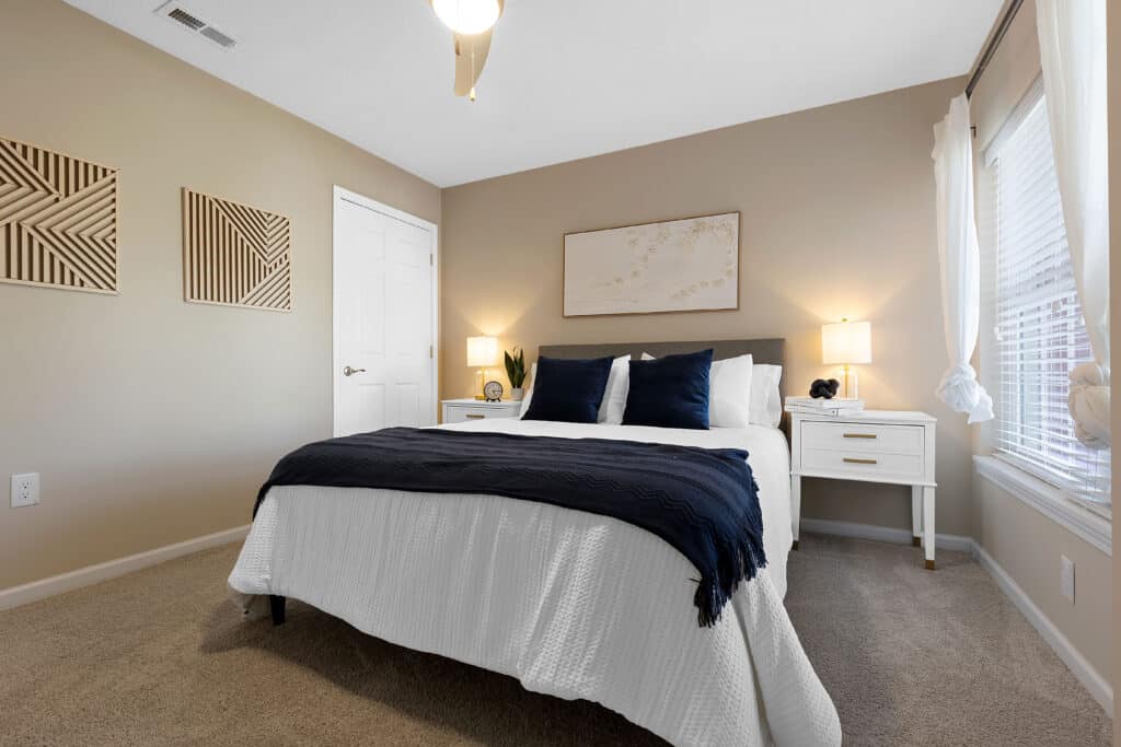 A neatly made bed with dark blue accents sits in a carpeted bedroom with beige walls, two nightstands, lamps, and a window with white curtains.