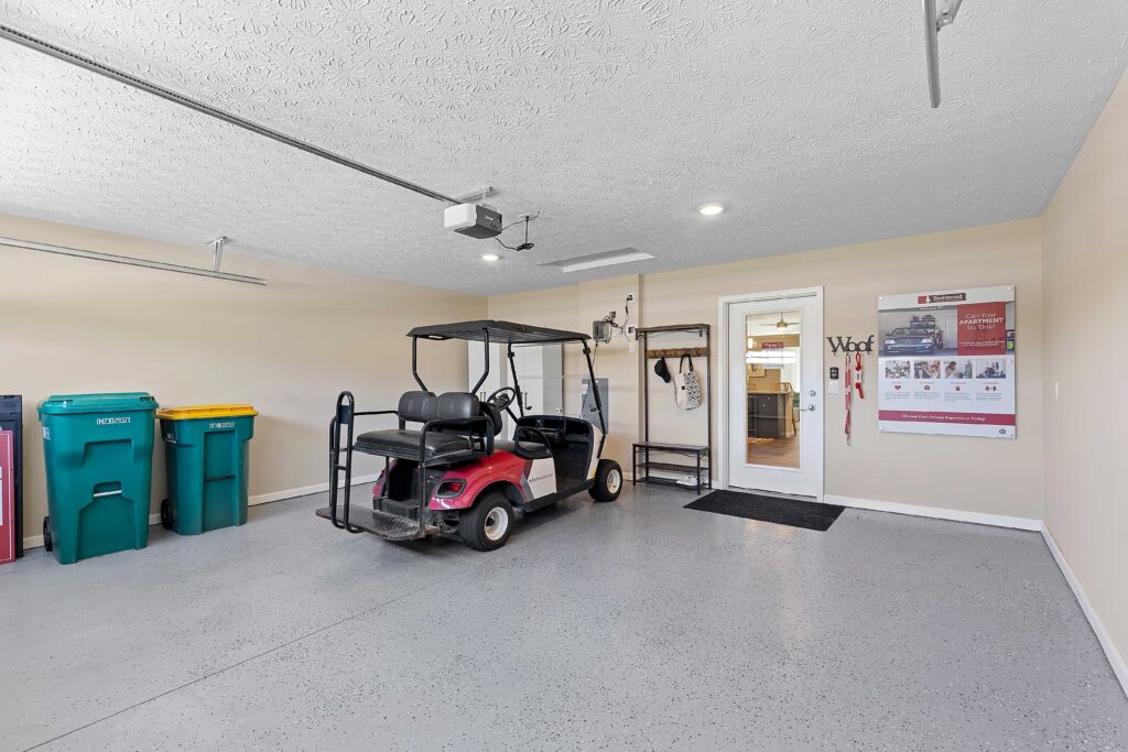 A pink golf cart is parked in a clean garage with recycling bins to the left and a white door leading inside. A wall calendar and shelves are mounted on the right wall.