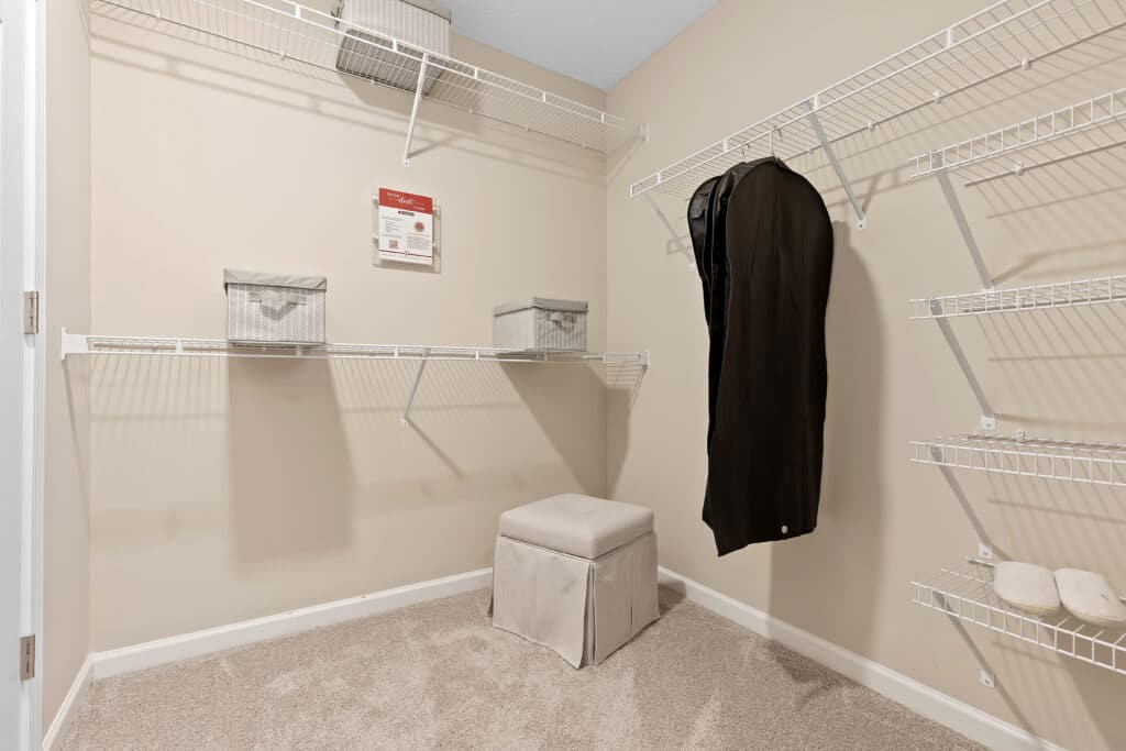 Empty walk-in closet with wire shelving, two gray storage boxes, a black garment bag hanging, a lidded beige ottoman, and beige carpeted floor.