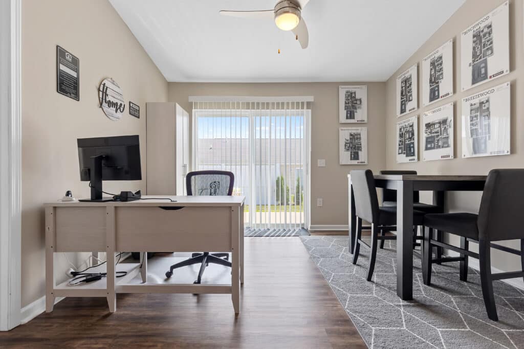 Modern home office with a desk, computer, and chair on the left, a small dining table with chairs on the right, and large windows with blinds in the background.