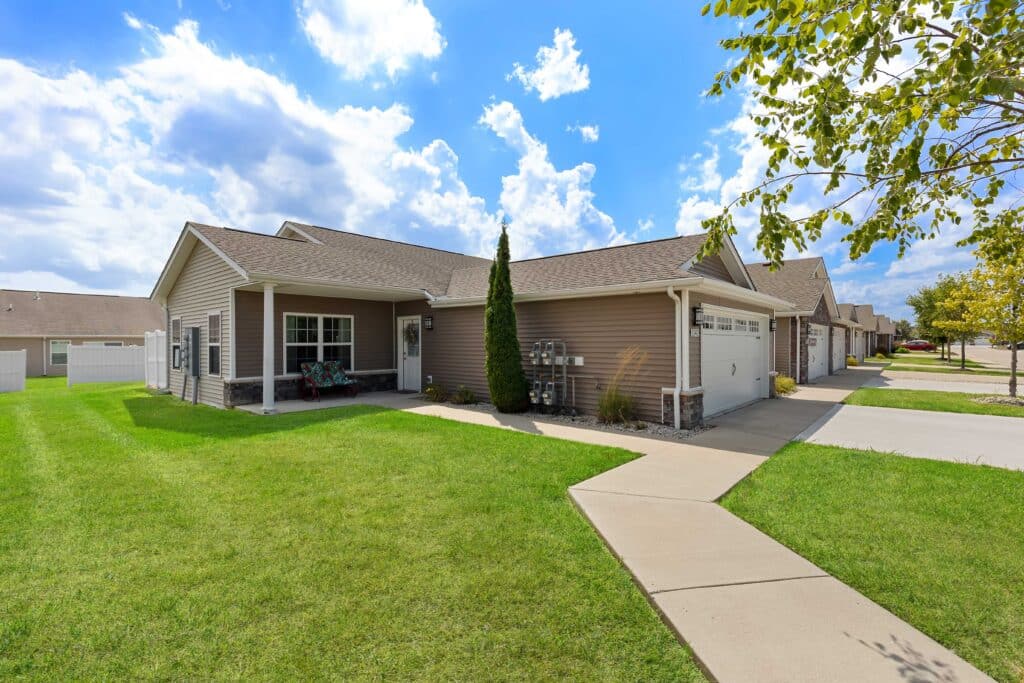 Single-story suburban townhouse with beige siding, two-car garage, manicured lawn, sidewalk, and partly cloudy sky.