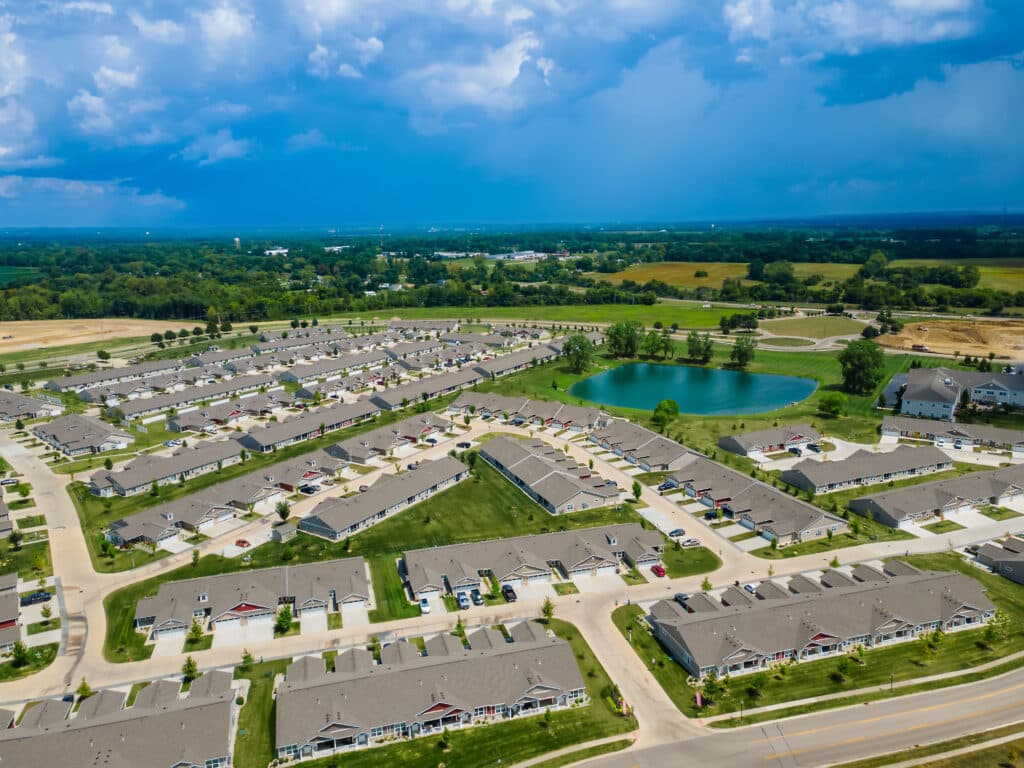 Aerial view of a suburban neighborhood with rows of single-story houses, green lawns, streets, and a pond surrounded by open fields under a partly cloudy sky.