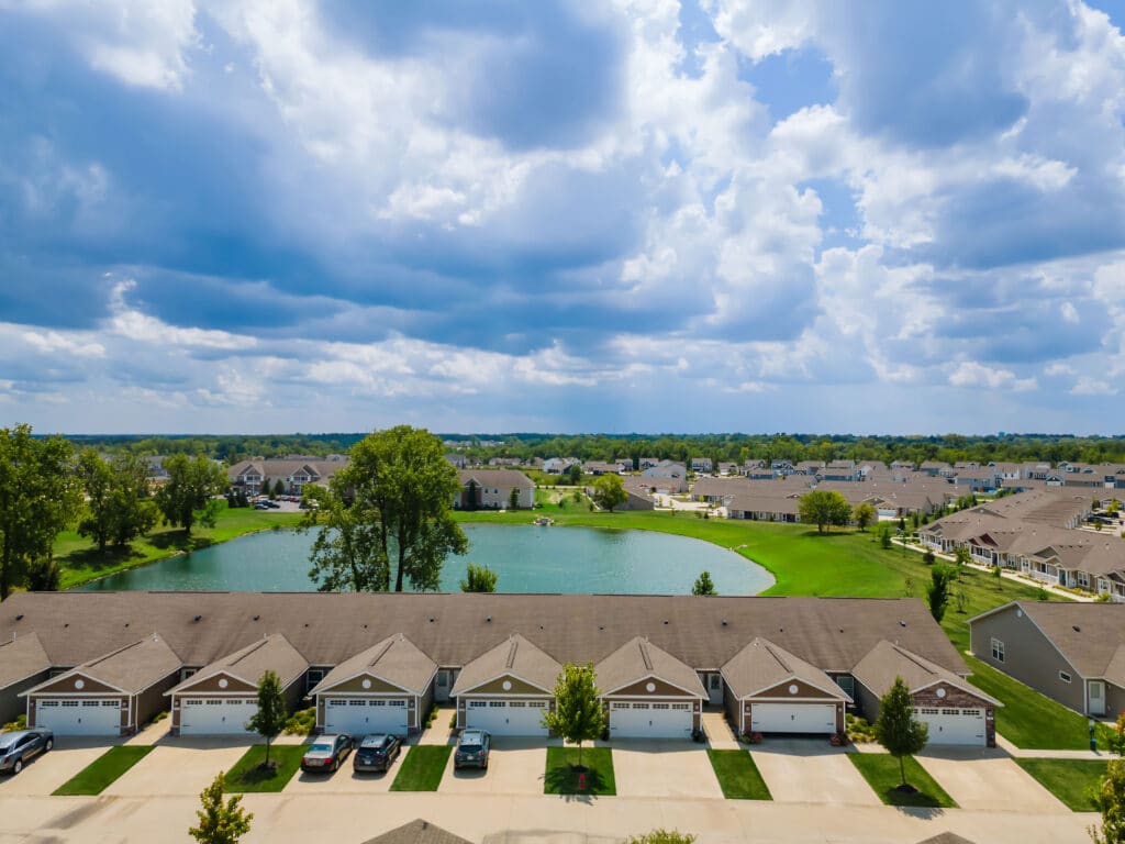 Aerial view of a suburban neighborhood with rows of houses, garages, green lawns, a pond, and trees under a partly cloudy sky.