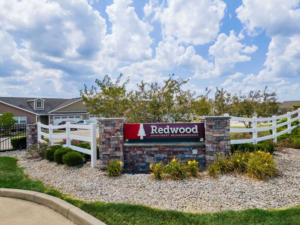 A stone sign reading "Redwood Apartment Neighborhoods" stands in landscaped gravel with shrubs, in front of white fencing and residential buildings under a partly cloudy sky.