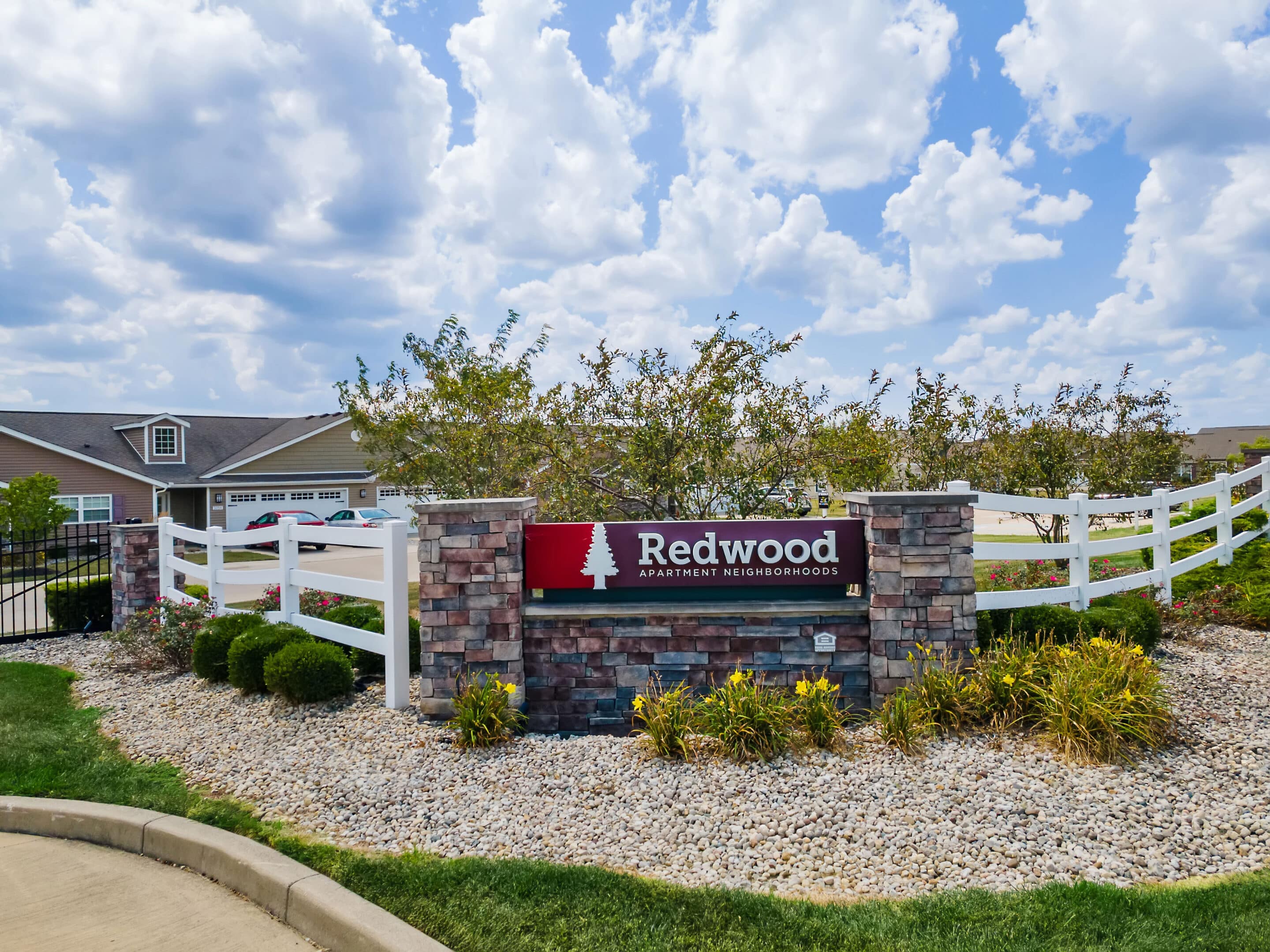 A stone sign reading "Redwood Apartment Neighborhoods" stands in landscaped gravel with shrubs, in front of white fencing and residential buildings under a partly cloudy sky.