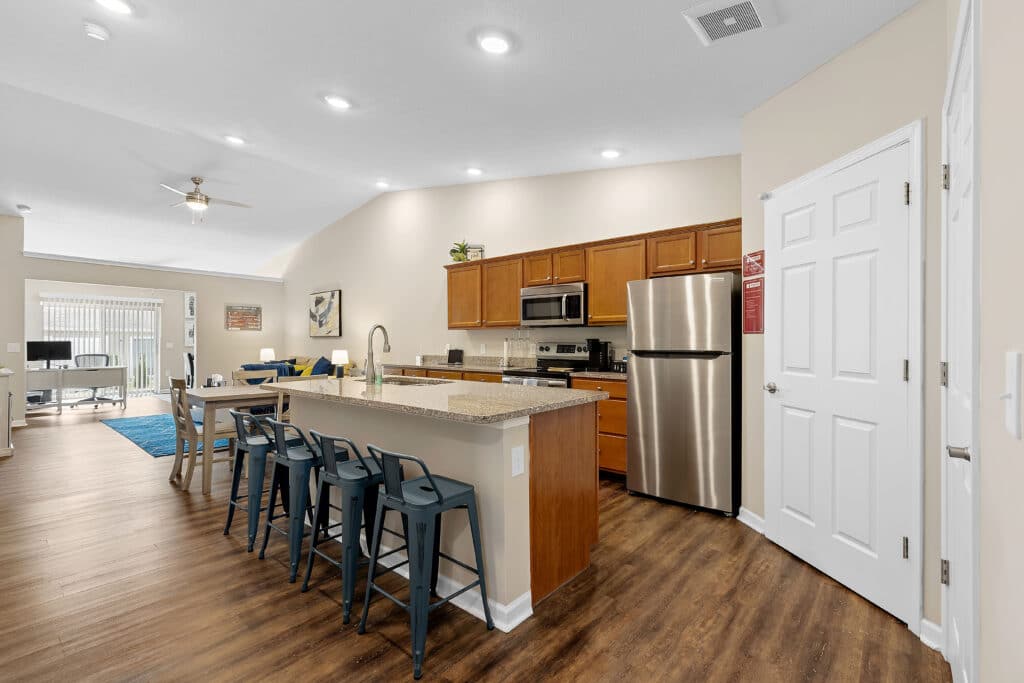 Modern kitchen with wooden cabinets, stainless steel appliances, granite island with bar stools, and open layout leading to a living area with a blue rug and large window.