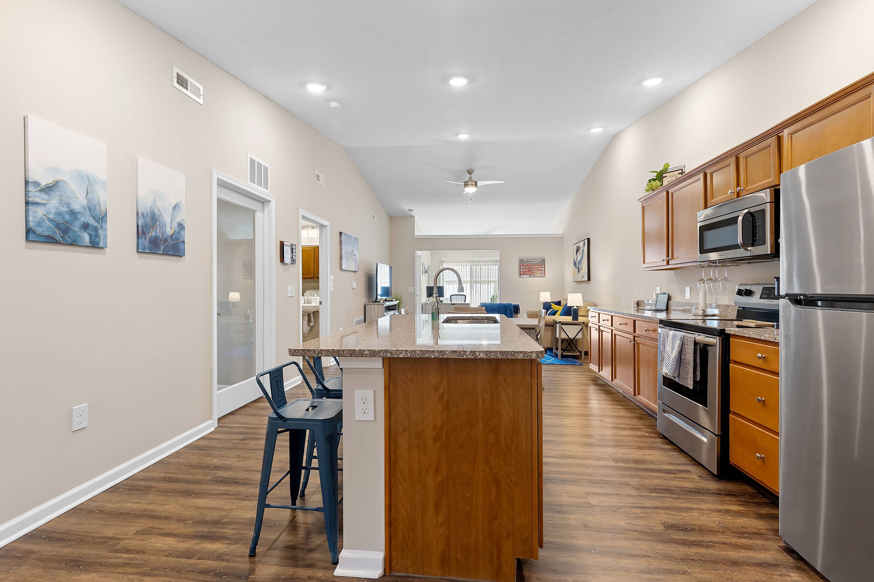Modern kitchen with stainless steel appliances, wooden cabinets, an island with two chairs, and an open layout leading to a living area with a large window.