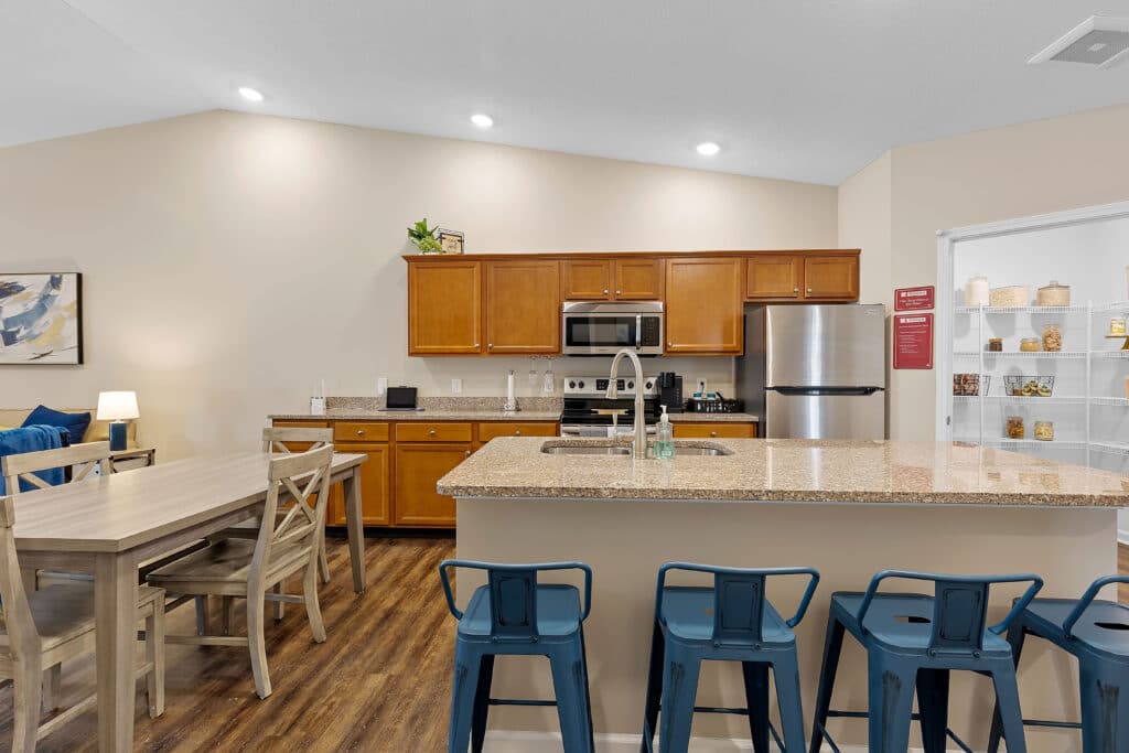 Modern kitchen with wooden cabinets, stainless steel appliances, granite countertops, blue barstools, dining table with chairs, and an open pantry stocked with jars and baskets.