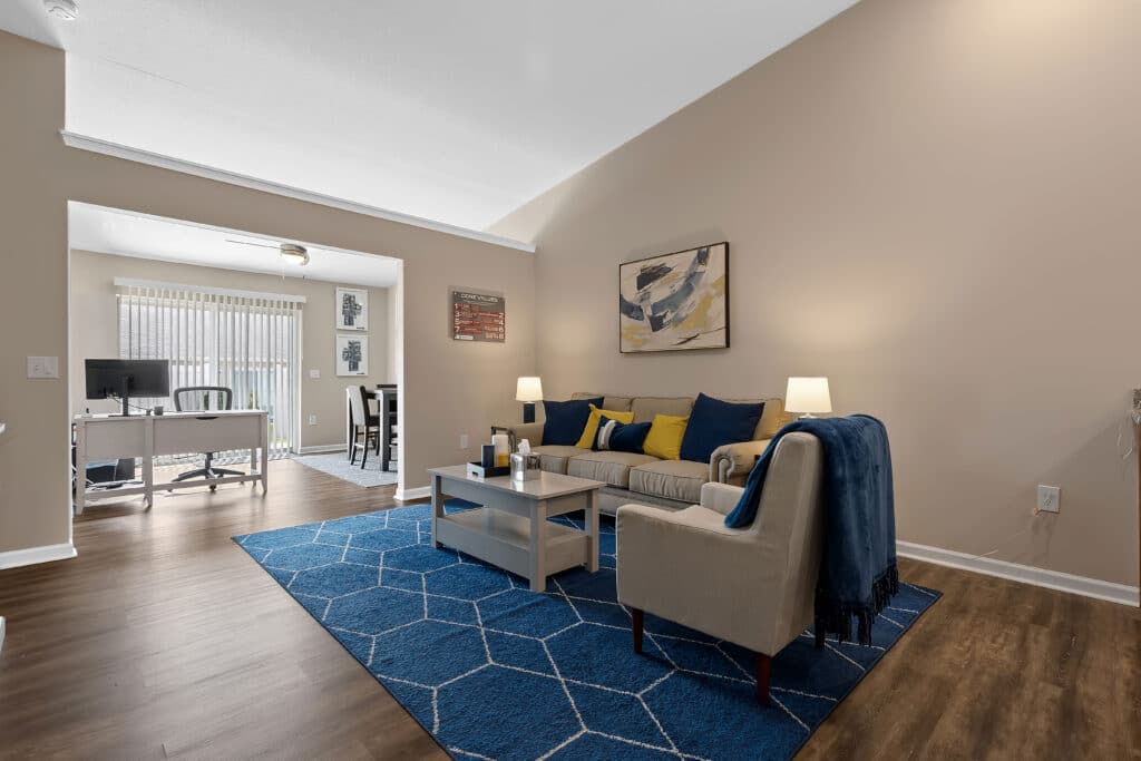 Modern living room with beige walls, blue rug, gray sofa, armchair, coffee table, and lamps; dining area and home office visible in the background through sliding glass doors.