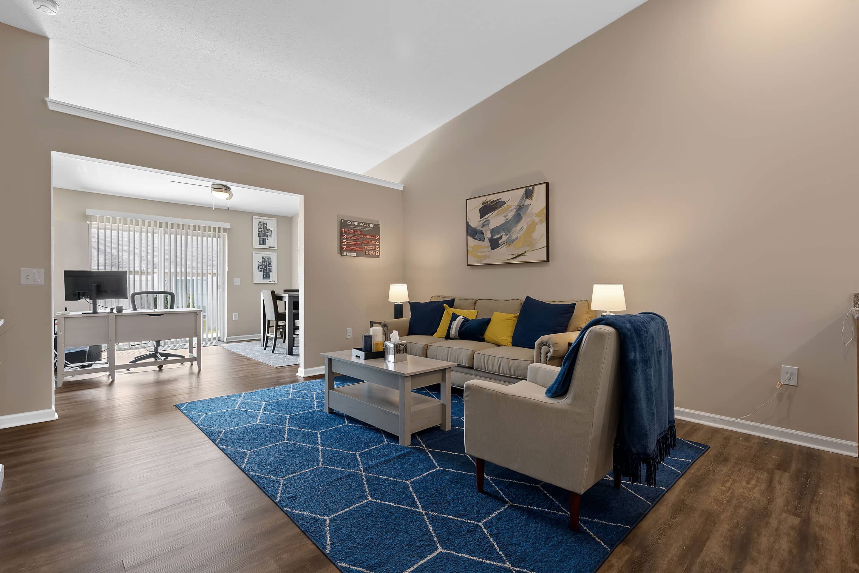 Modern living room with beige walls, blue rug, gray sofa, armchair, coffee table, and lamps; dining area and home office visible in the background through sliding glass doors.