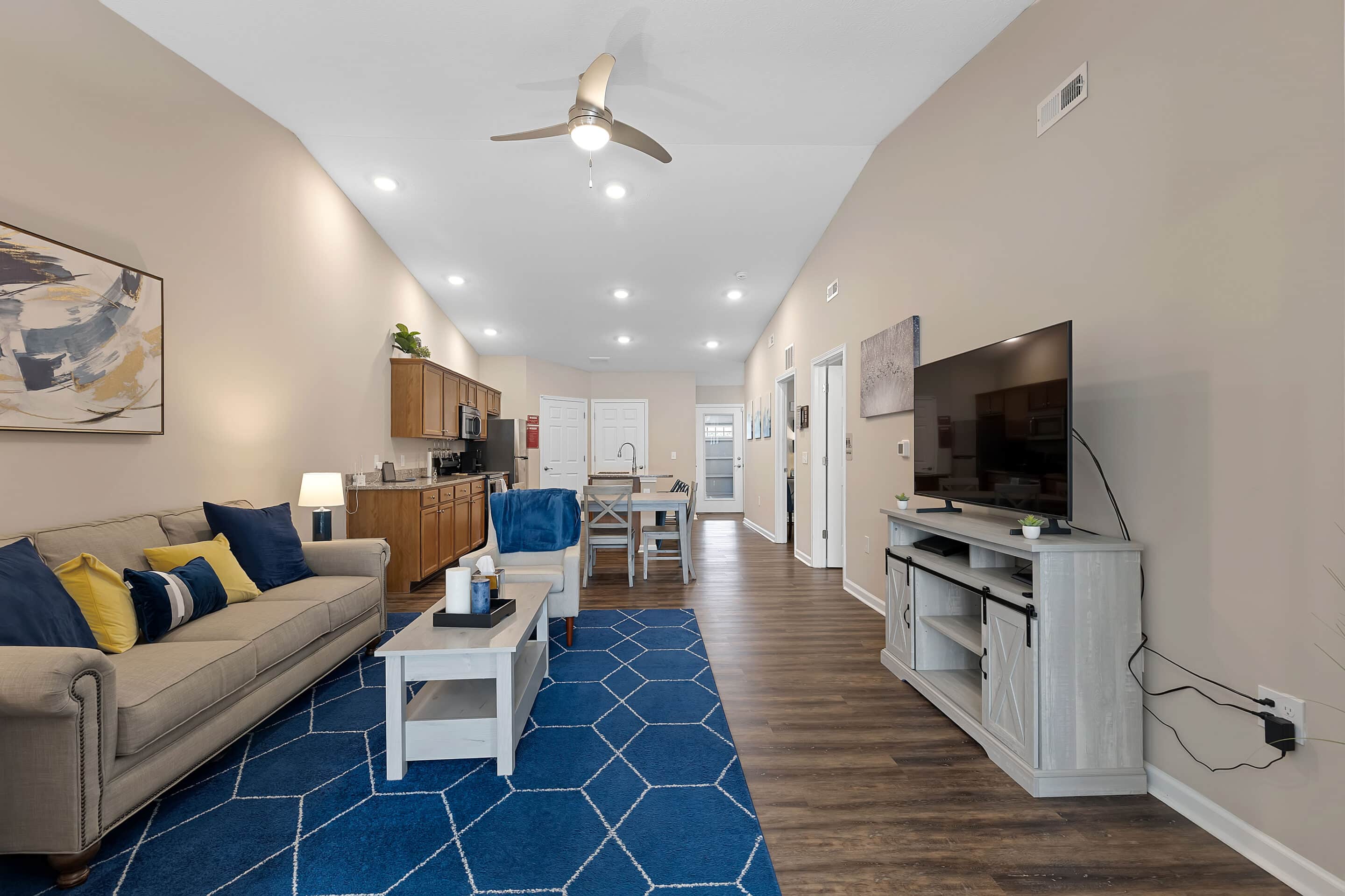 Open-concept living room and kitchen with beige walls, wood flooring, a gray sofa, blue rug, TV on a stand, dining table, and recessed lighting.