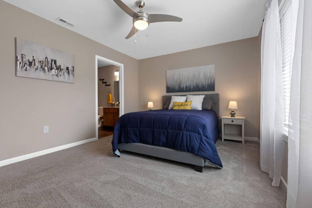 A modern bedroom with a gray bed frame, blue bedding, two nightstands with lamps, wall art, carpeted floor, and a ceiling fan. An open door leads to a bathroom.