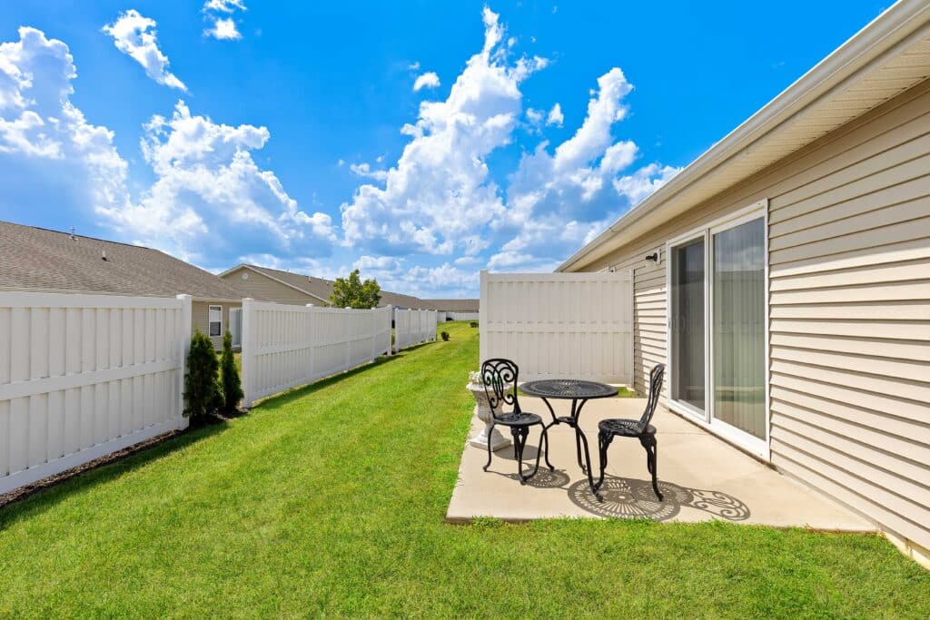 A small concrete patio with a black metal table and two chairs sits next to a beige house, bordered by a white fence and green lawn under a blue sky with clouds.