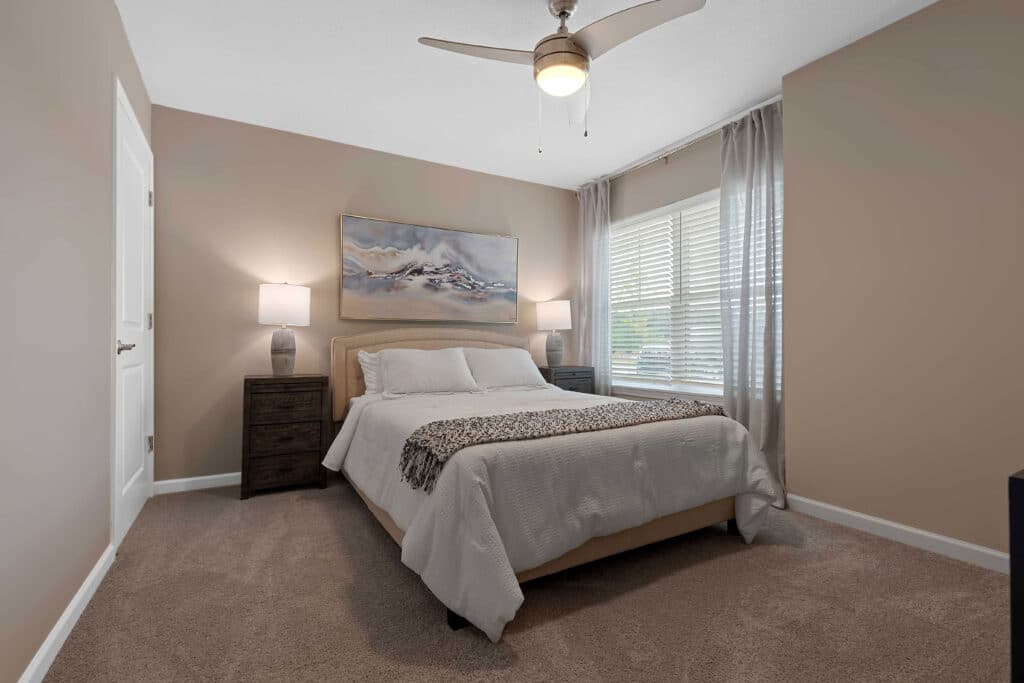 A neatly made bed with white bedding, two dark nightstands with lamps, a ceiling fan, artwork above the bed, and a window with sheer curtains in a beige bedroom.