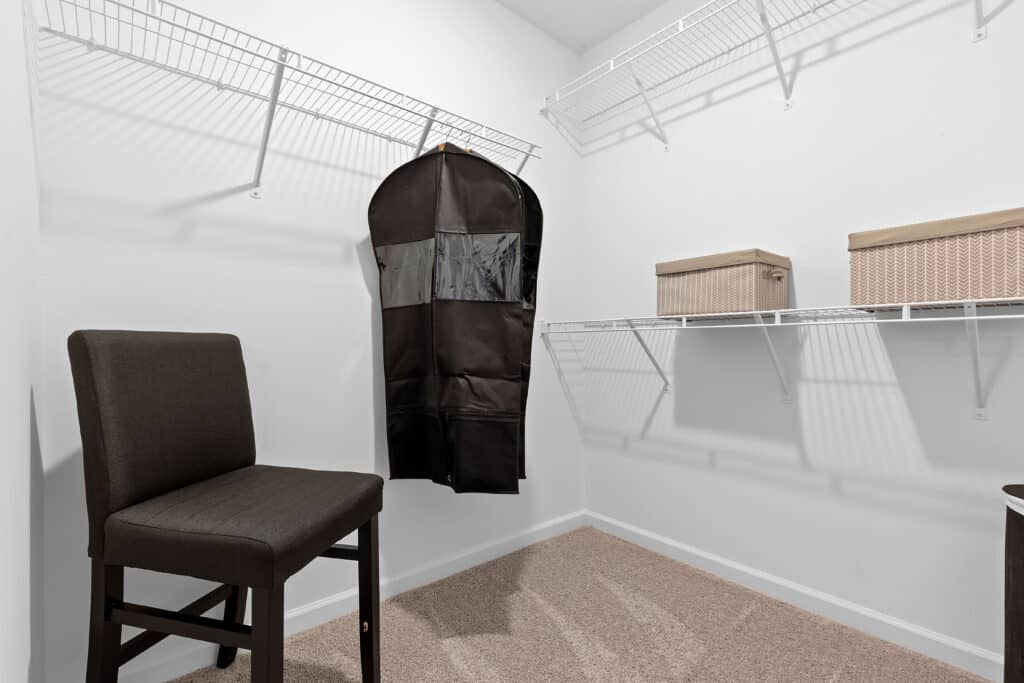 A walk-in closet with white walls, wire shelves, a brown chair, a hanging garment bag, and two woven storage boxes on the shelves.