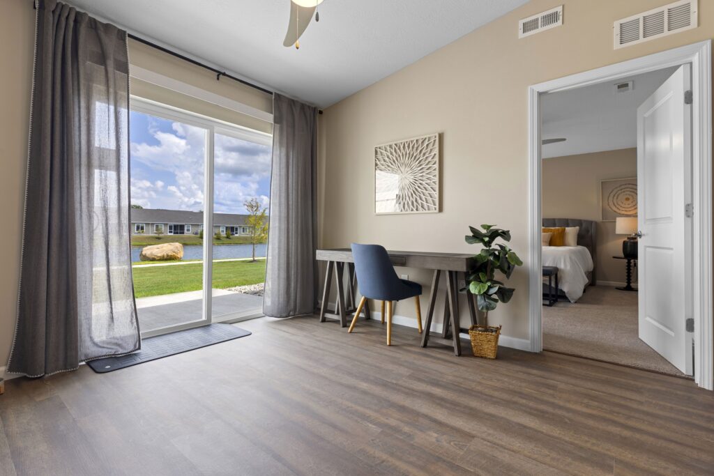A home office with a desk and chair, potted plant, and large window with sheer curtains overlooking a lake. An open door leads to a bedroom with a bed and wall art.