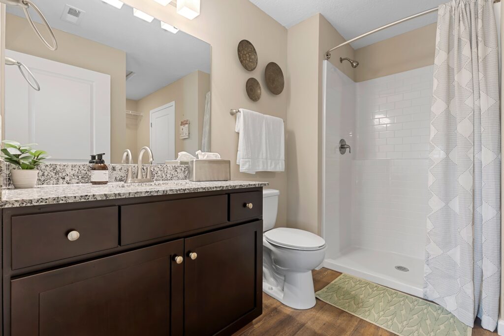 Modern bathroom with a dark wood vanity, granite countertop, toilet, and white-tiled shower with a curtain. Towels, plants, and decorative plates add simple accents.
