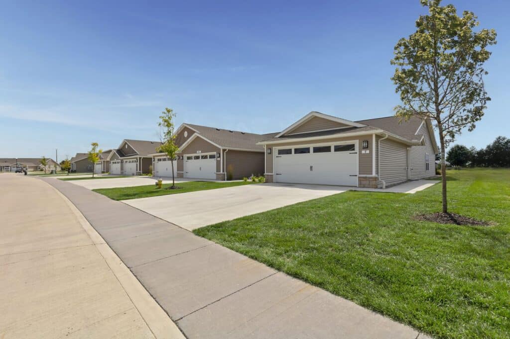 Row of modern suburban houses with attached garages, neatly trimmed lawns, and young trees lining a residential street under a clear blue sky.